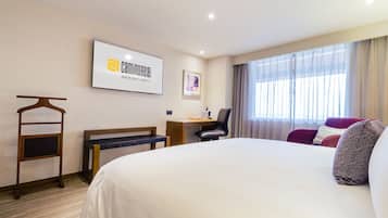 Habitación de hotel con una cama grande, un escritorio con silla, un televisor montado en la pared y un ventanal con cortinas.