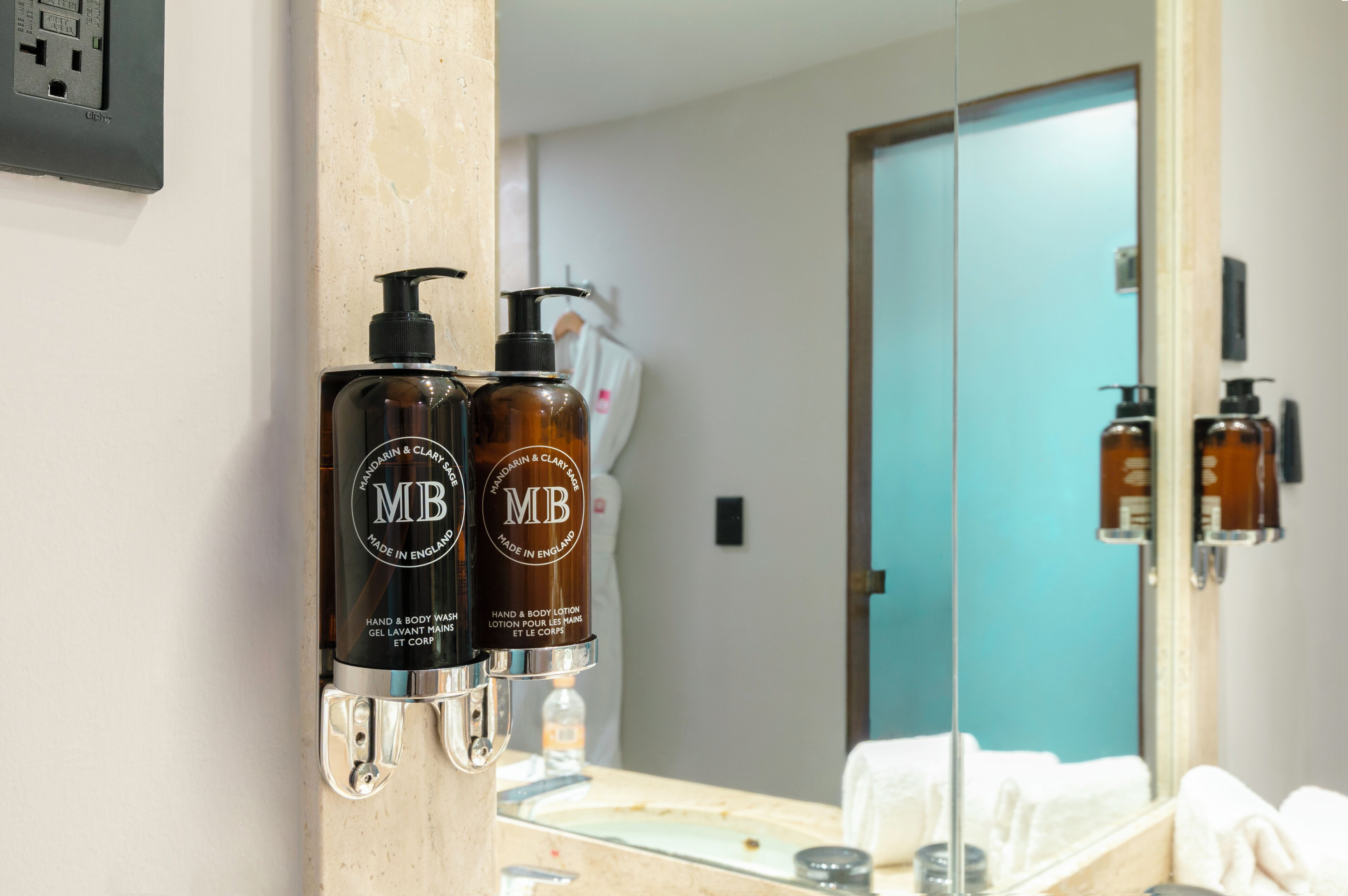 A bathroom with two hand and body lotion dispensers labeled 'MB' on a beige countertop, a mirror reflecting the room, and a door in the background.