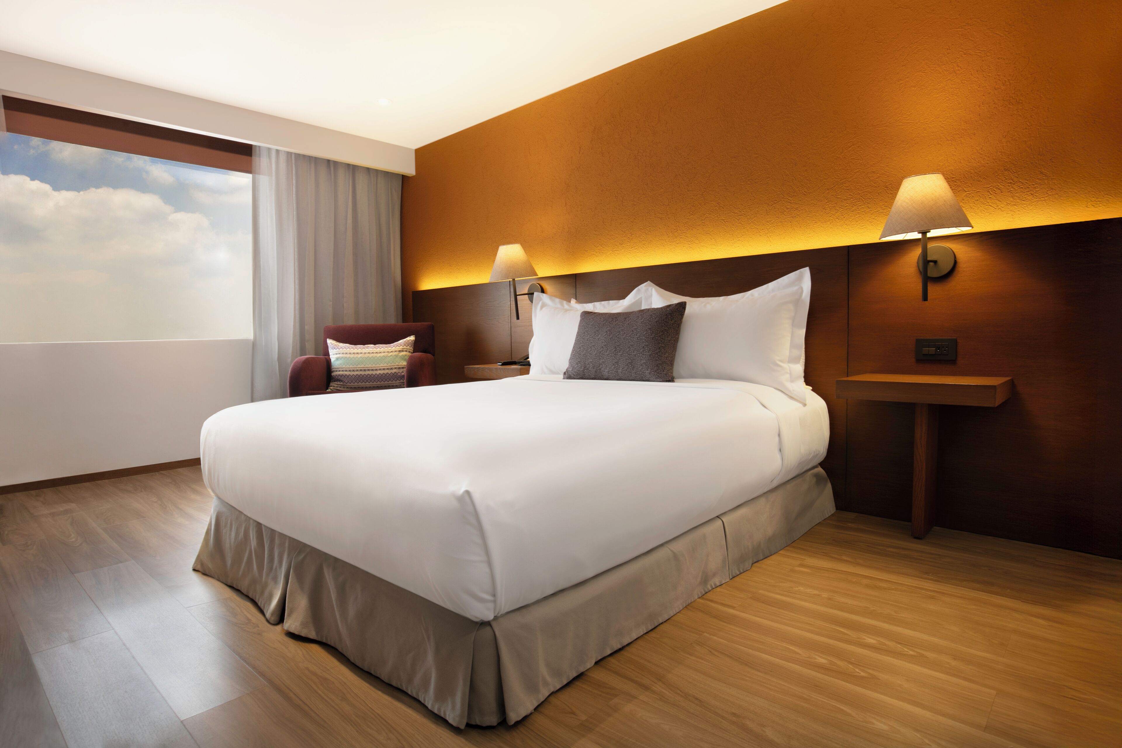 Une chambre d’hôtel avec un grand lit, deux tables de chevet, une chaise et une vue sur le ciel par une fenêtre.