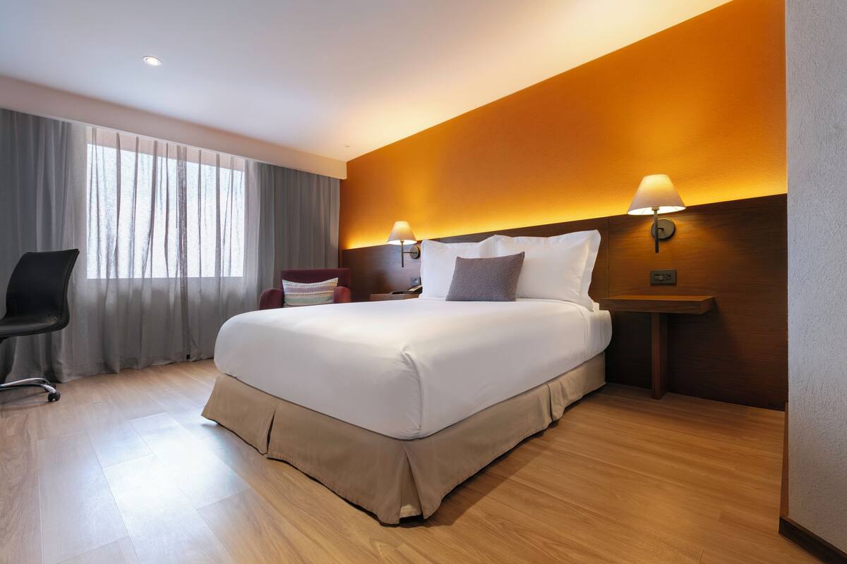 A hotel room with a large bed, two bedside lamps, a chair, and a window with curtains.