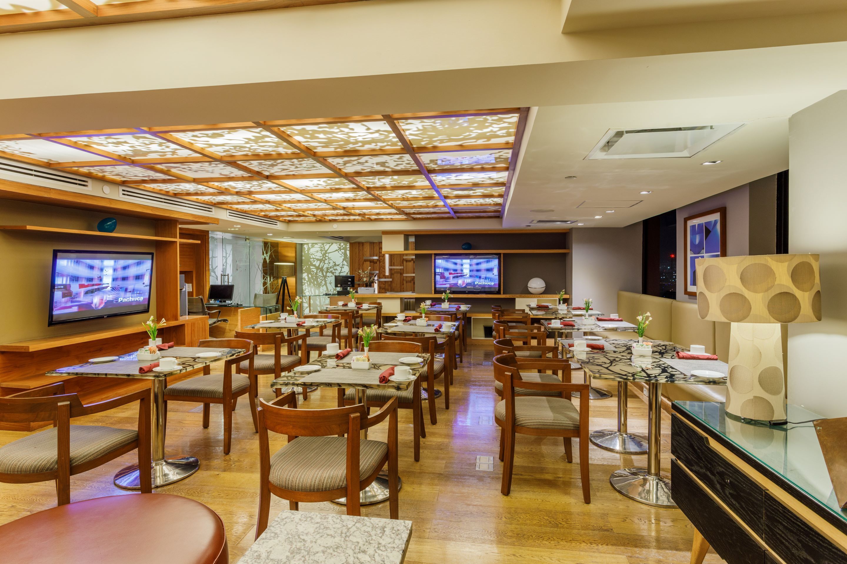 Een modern restaurant met meerdere eettafels en stoelen, een plafond met een geometrisch patroon, en twee televisies aan de muur.