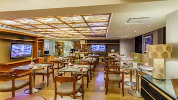 Een modern restaurant met meerdere eettafels en stoelen, een plafond met een geometrisch patroon, en twee televisies aan de muur.