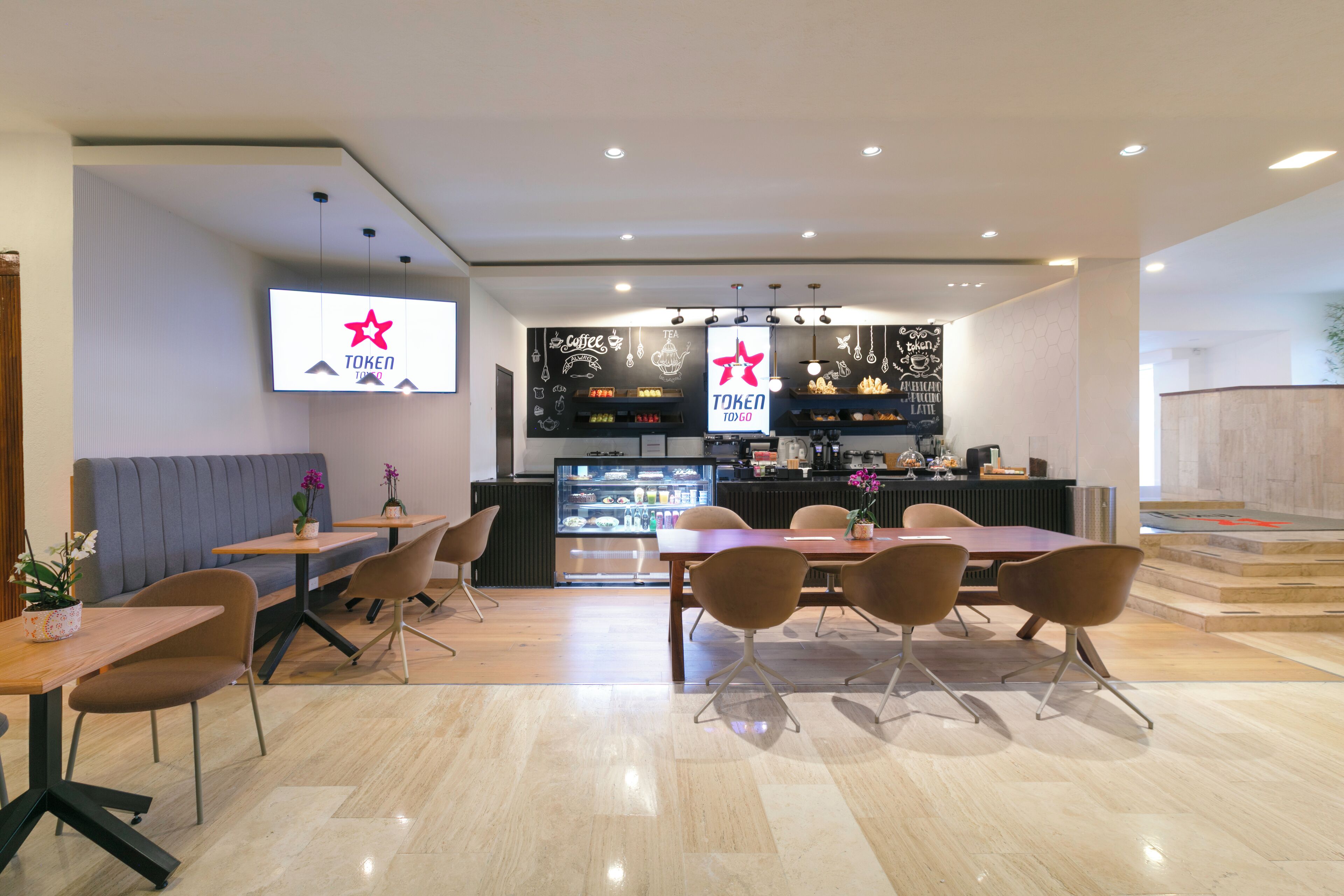 Un café moderno con área de bar, diferentes disposiciones de asientos y una pantalla digital.