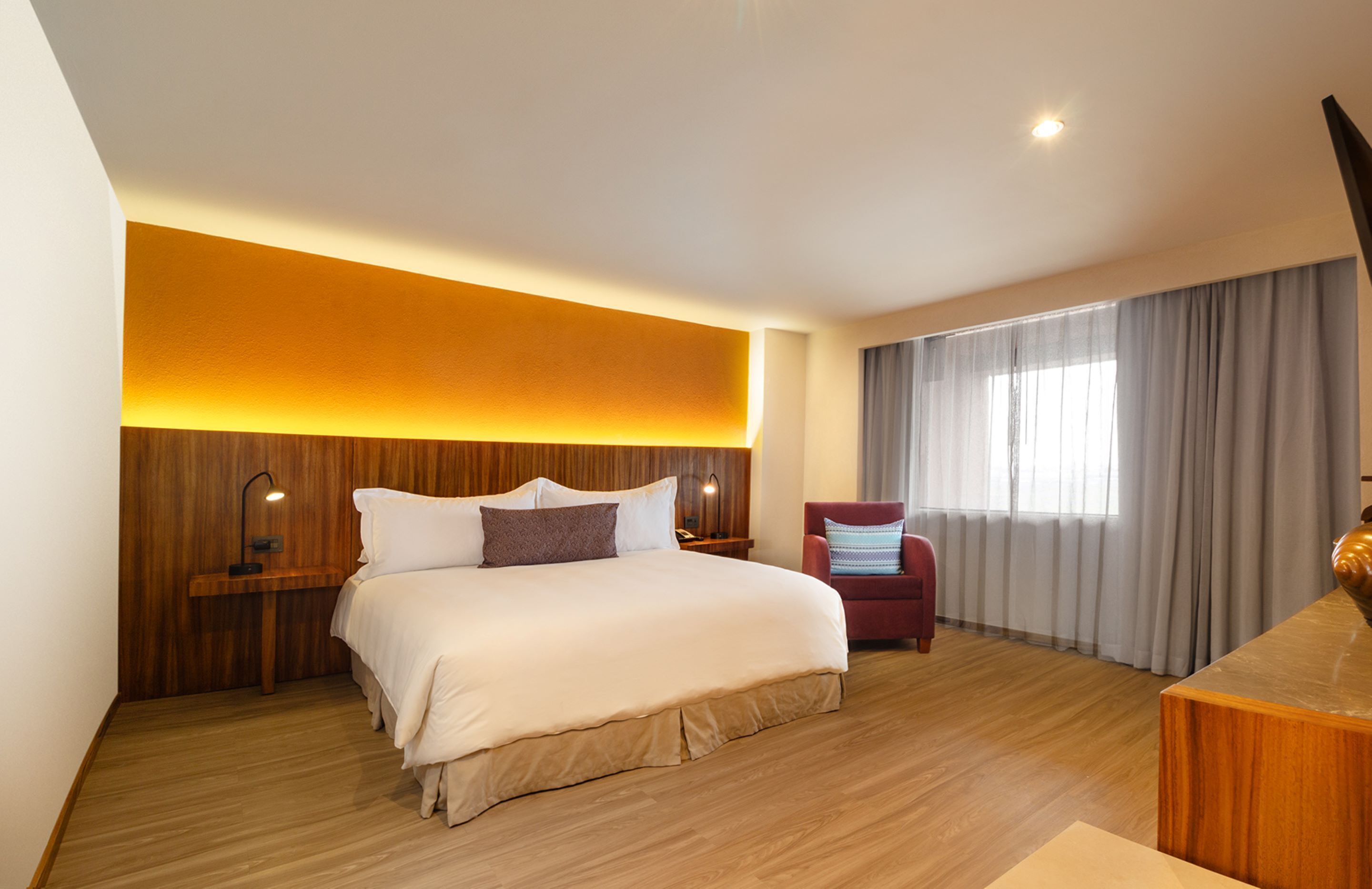 Una habitación de hotel moderna con una cama grande, cabecero de madera, lámparas de noche, un sillón rojo y un ventanal con cortinas.