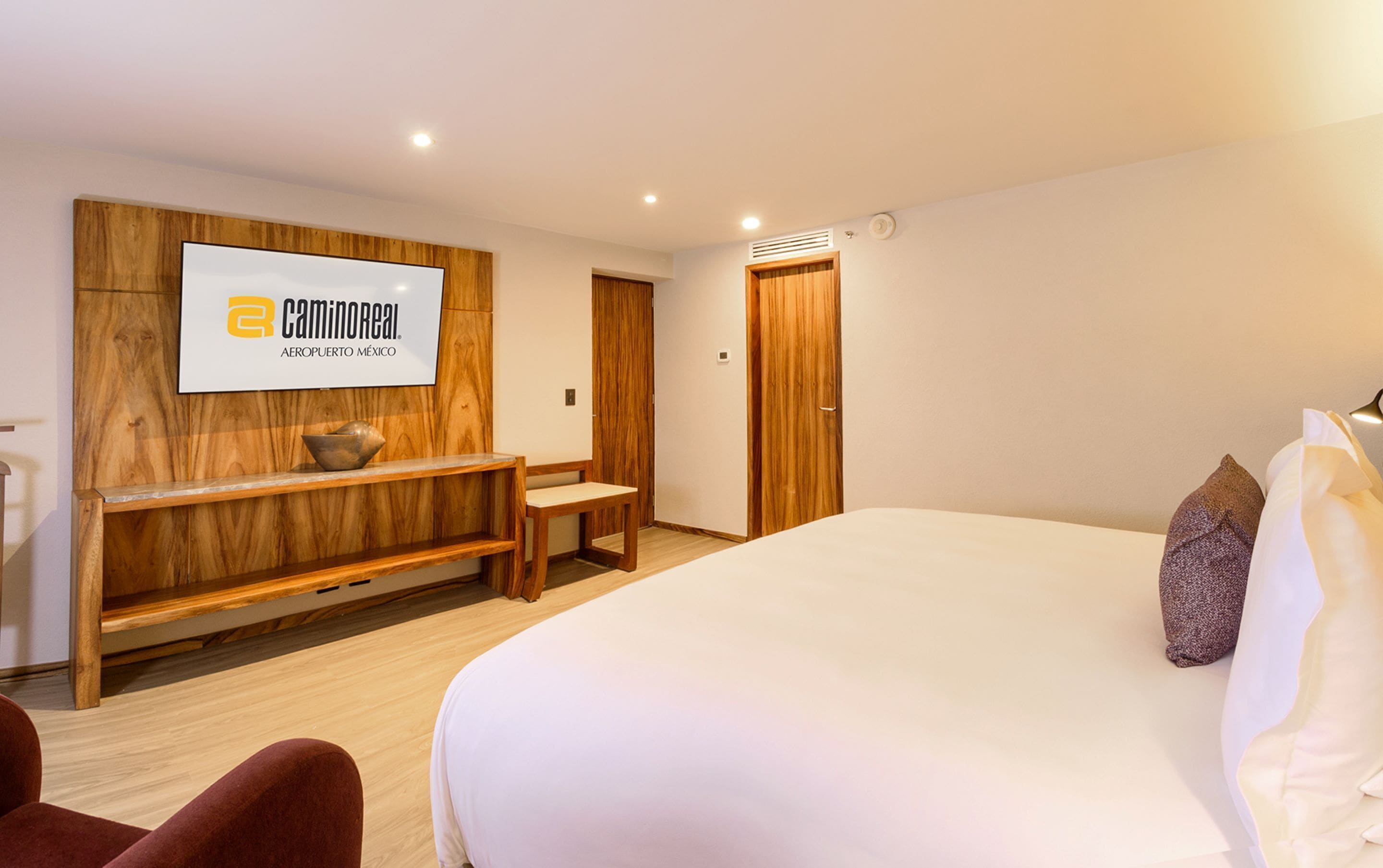 A hotel room with a large bed, a wooden bench, a TV mounted on a wooden panel, and a door with a window.