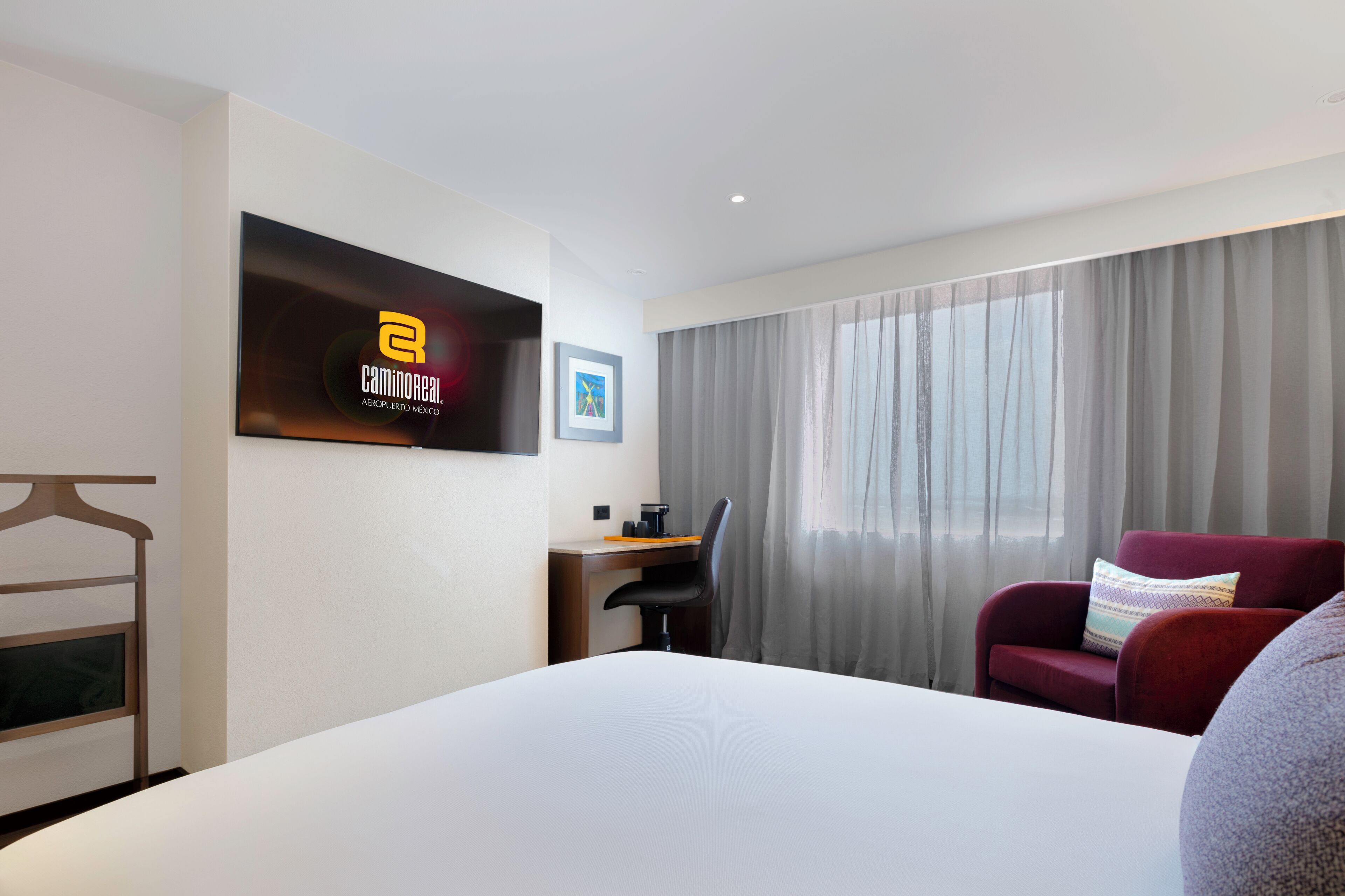 A hotel room with a bed, a desk with a chair, a red armchair, a television mounted on the wall, and a window with curtains.