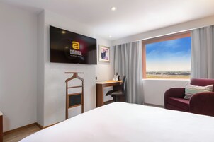 A hotel room with a bed, a desk, a chair, a TV mounted on the wall, and a window with a view.