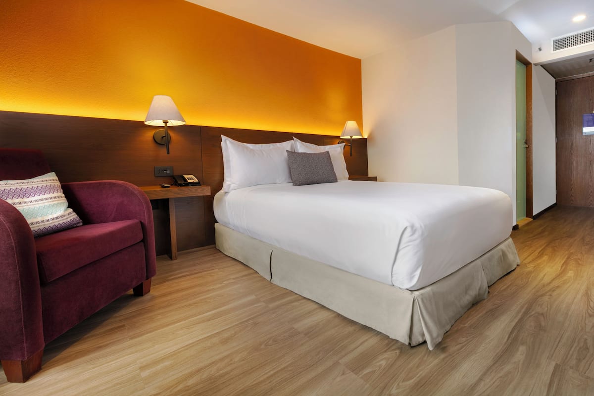 A hotel room with a large bed, a red armchair, two bedside lamps, and a wooden floor.