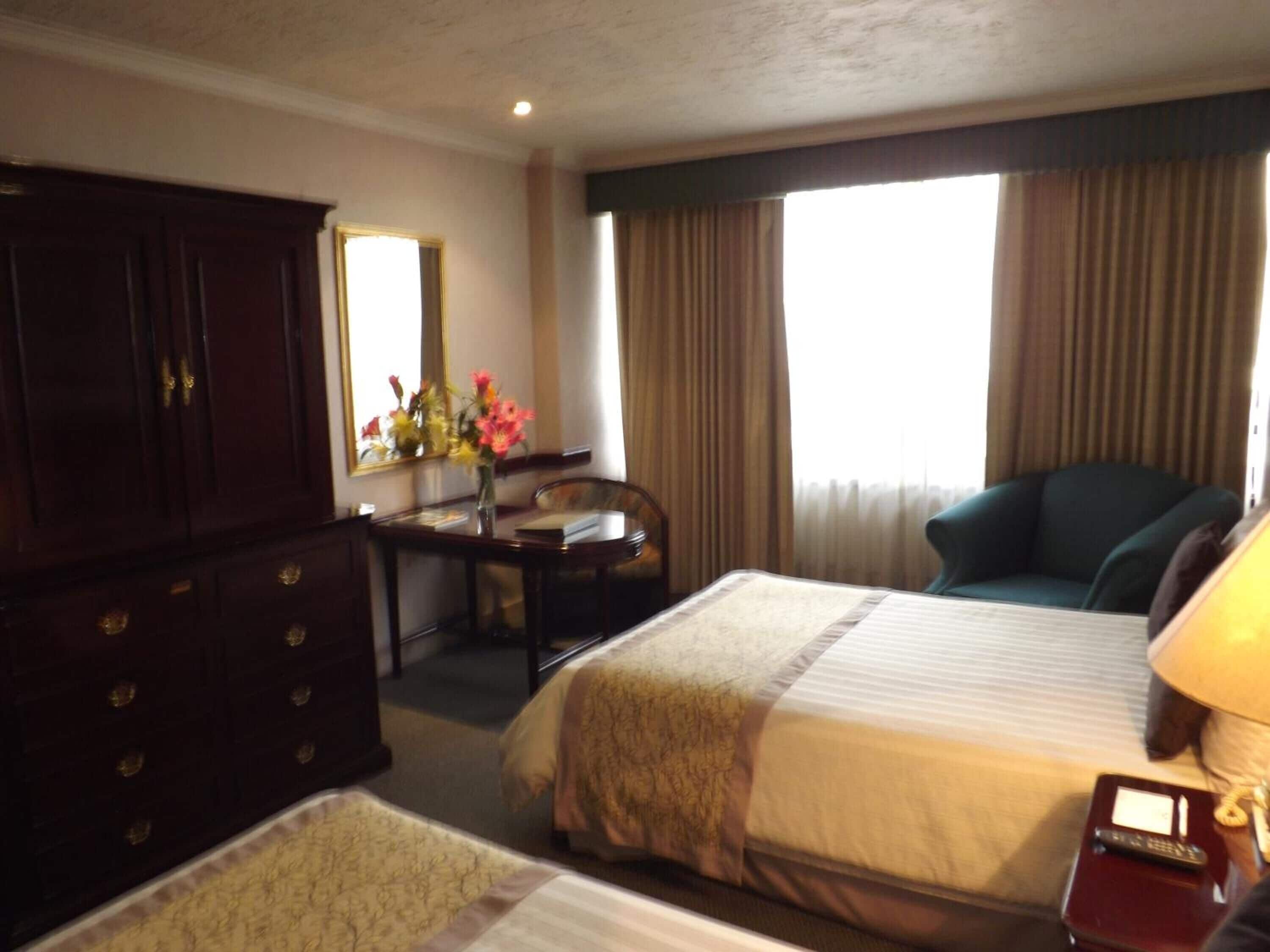 Une chambre d’hôtel avec deux lits, un bureau, une chaise, une commode et une grande fenêtre avec des rideaux.