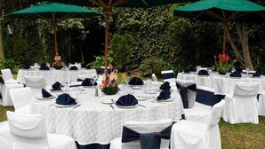 An outdoor dining area with round tables covered in white tablecloths, navy blue chair covers, and green umbrellas providing shade.