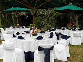 Un espace de restauration en plein air avec des tables rondes recouvertes de nappes blanches, des housses de chaise bleu marine et des parasols verts offrant de l’ombre.