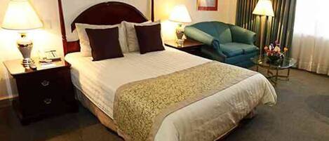 Habitación de hotel con una cama grande, dos mesitas de noche con lámparas, una silla y una pequeña mesa con flores.