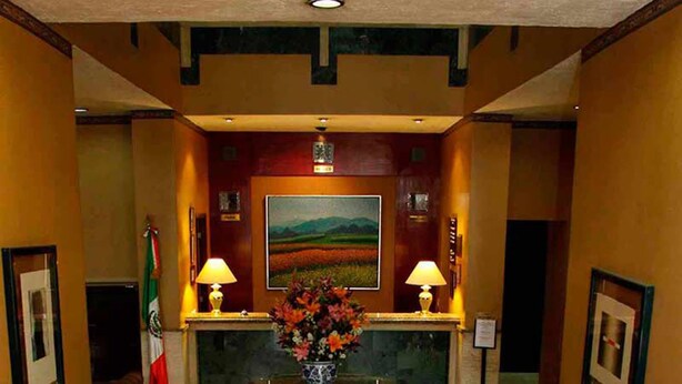 A lobby with a reception desk, two lamps, a vase of flowers, and four armchairs arranged around a central table.