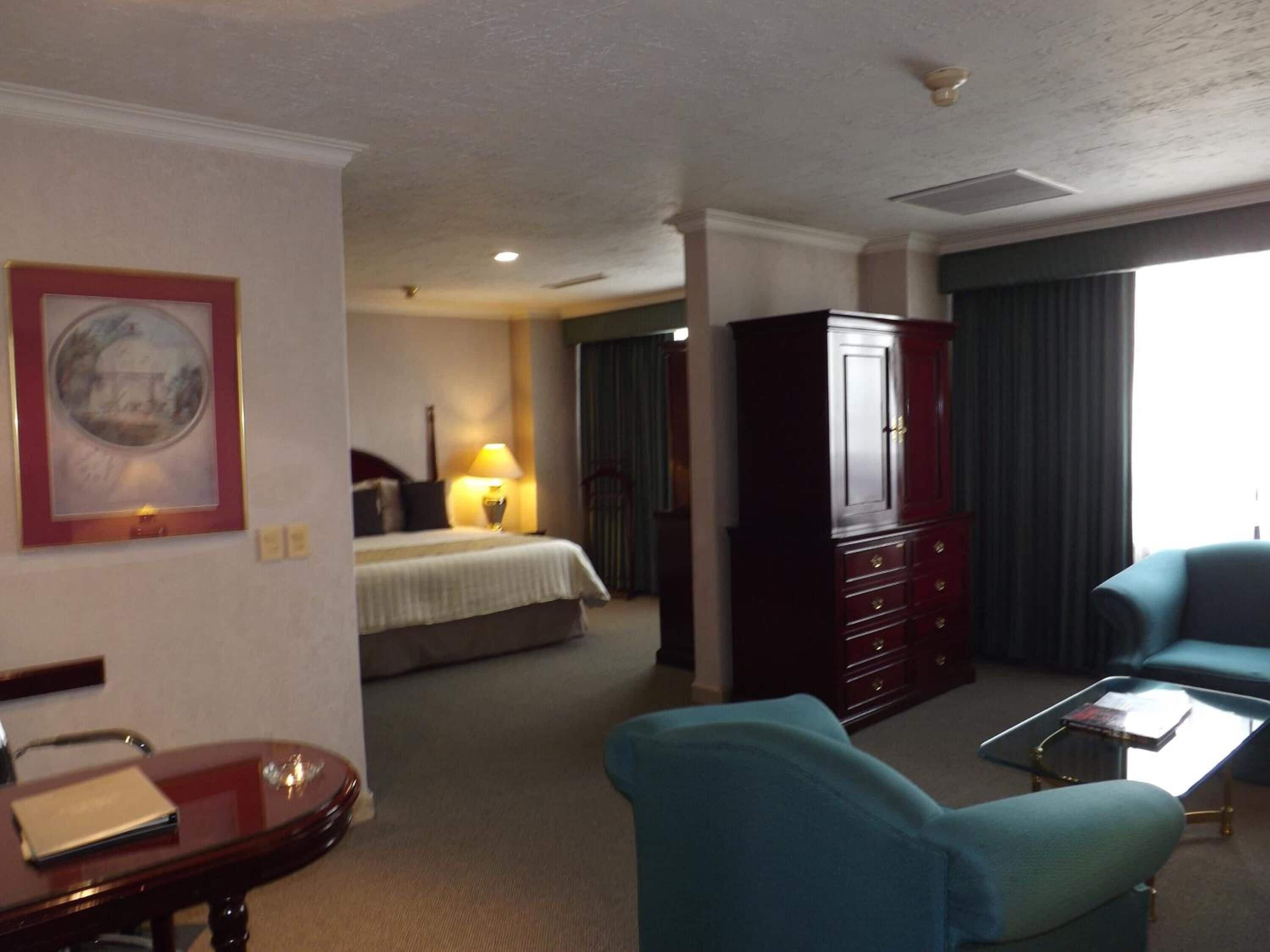 Una habitación de hotel con cama, escritorio, silla, armario y un ventanal con cortinas.