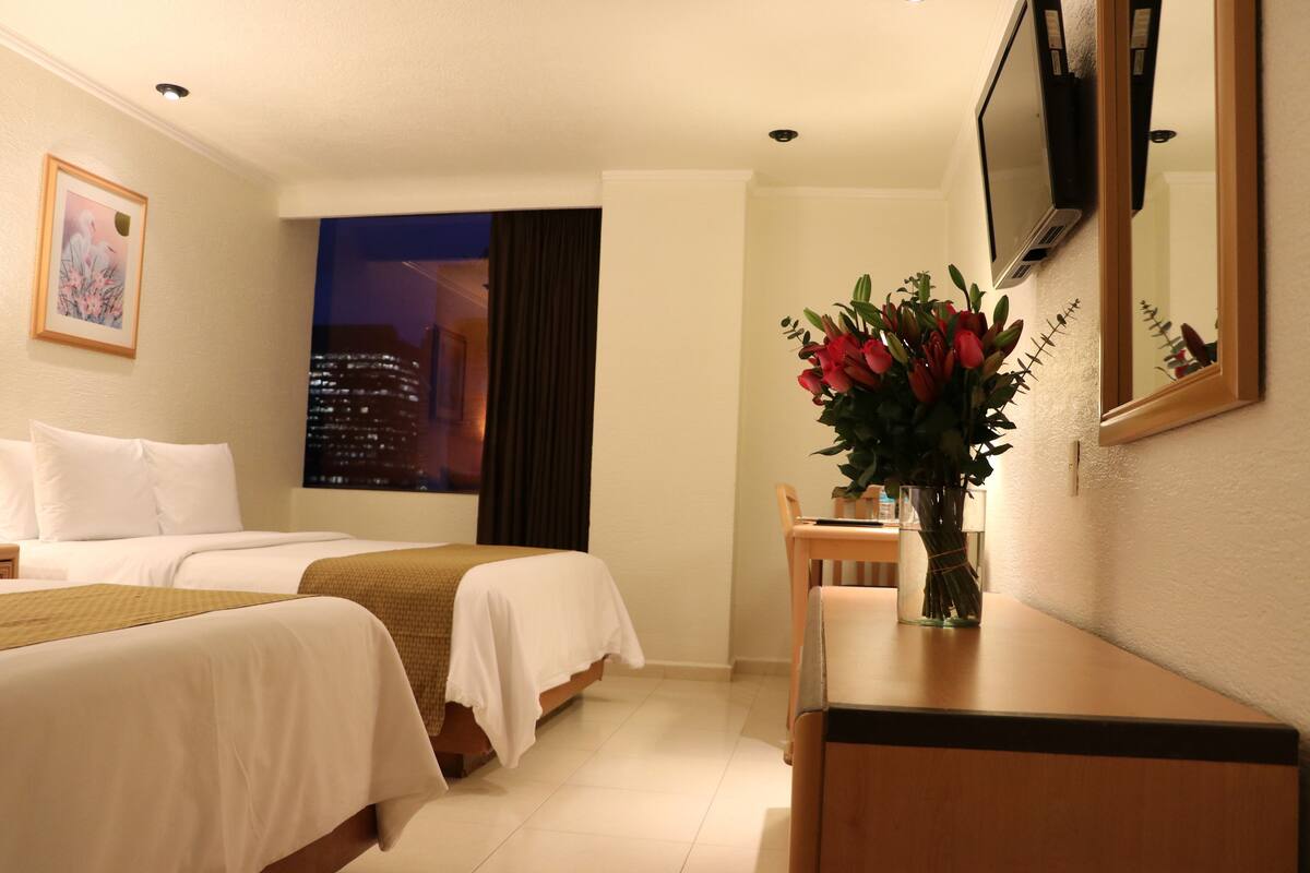Ett hotellrum med två sängar, ett skrivbord med en vas med blommor, utsikt över staden genom fönstret och en väggmonterad TV.