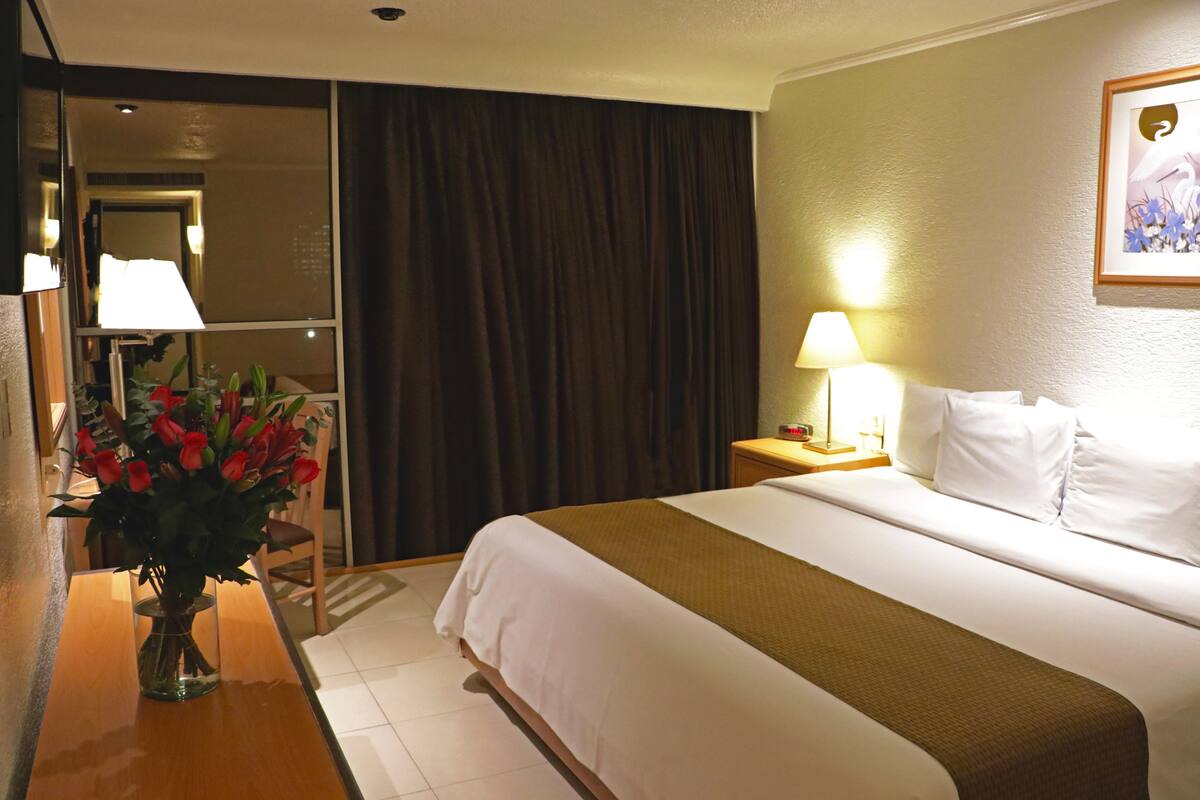 Ett hotellrum med en säng, ett nattduksbord, en lampa, en vas med blommor, en spegel och en gardin.