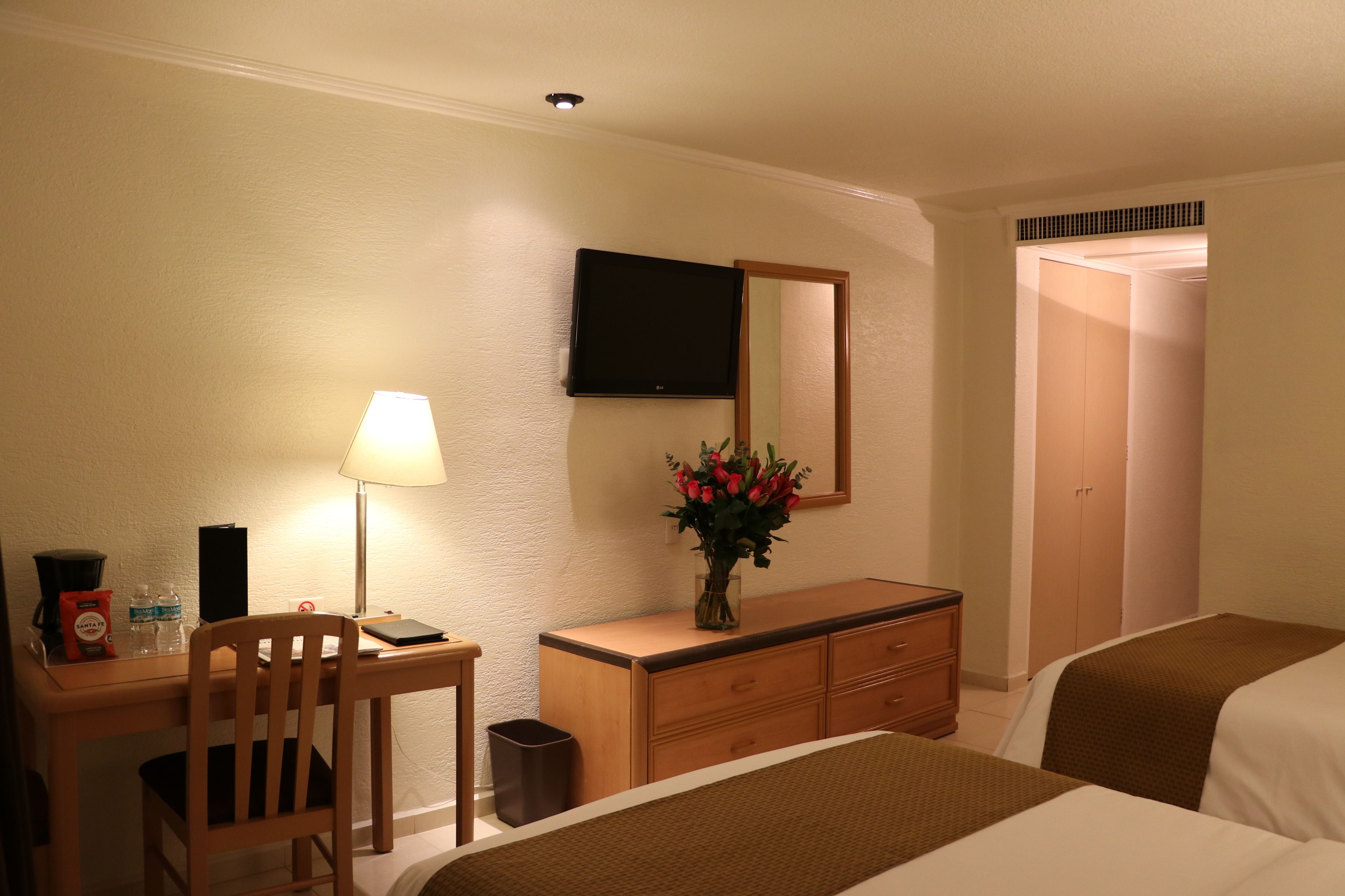 Habitación de hotel con cama, escritorio con lámpara, cómoda, televisor montado en la pared y espejo.