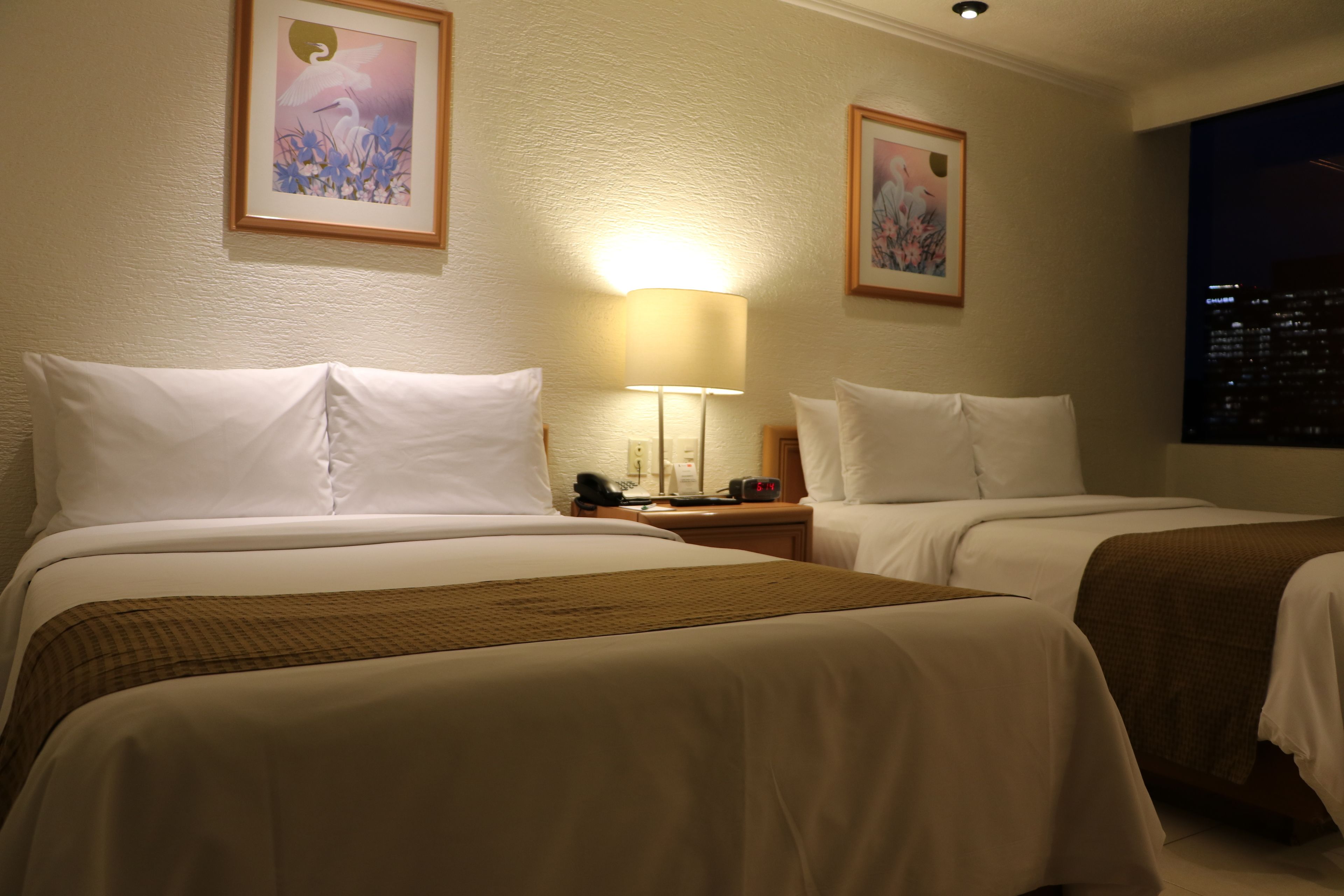 A hotel room with two beds, a nightstand with a lamp, and framed artwork on the walls.
