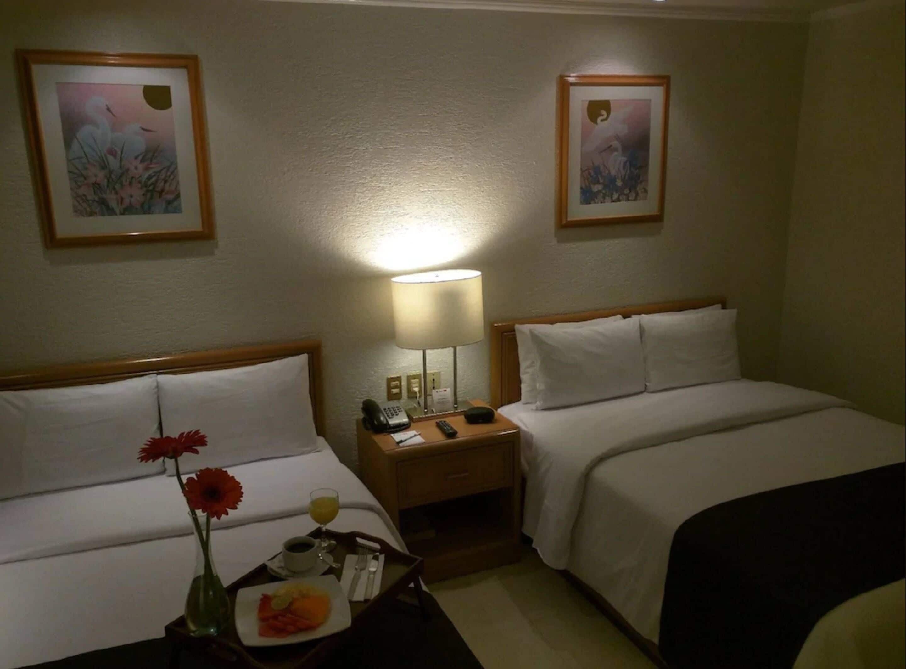 Une chambre d’hôtel avec deux lits, une table de chevet avec une lampe, un plateau avec des articles pour le petit-déjeuner et deux tableaux encadrés au mur.