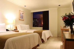Une chambre d’hôtel avec deux lits, un bureau et une chaise. Il y a une fenêtre qui offre une vue sur la ville la nuit.