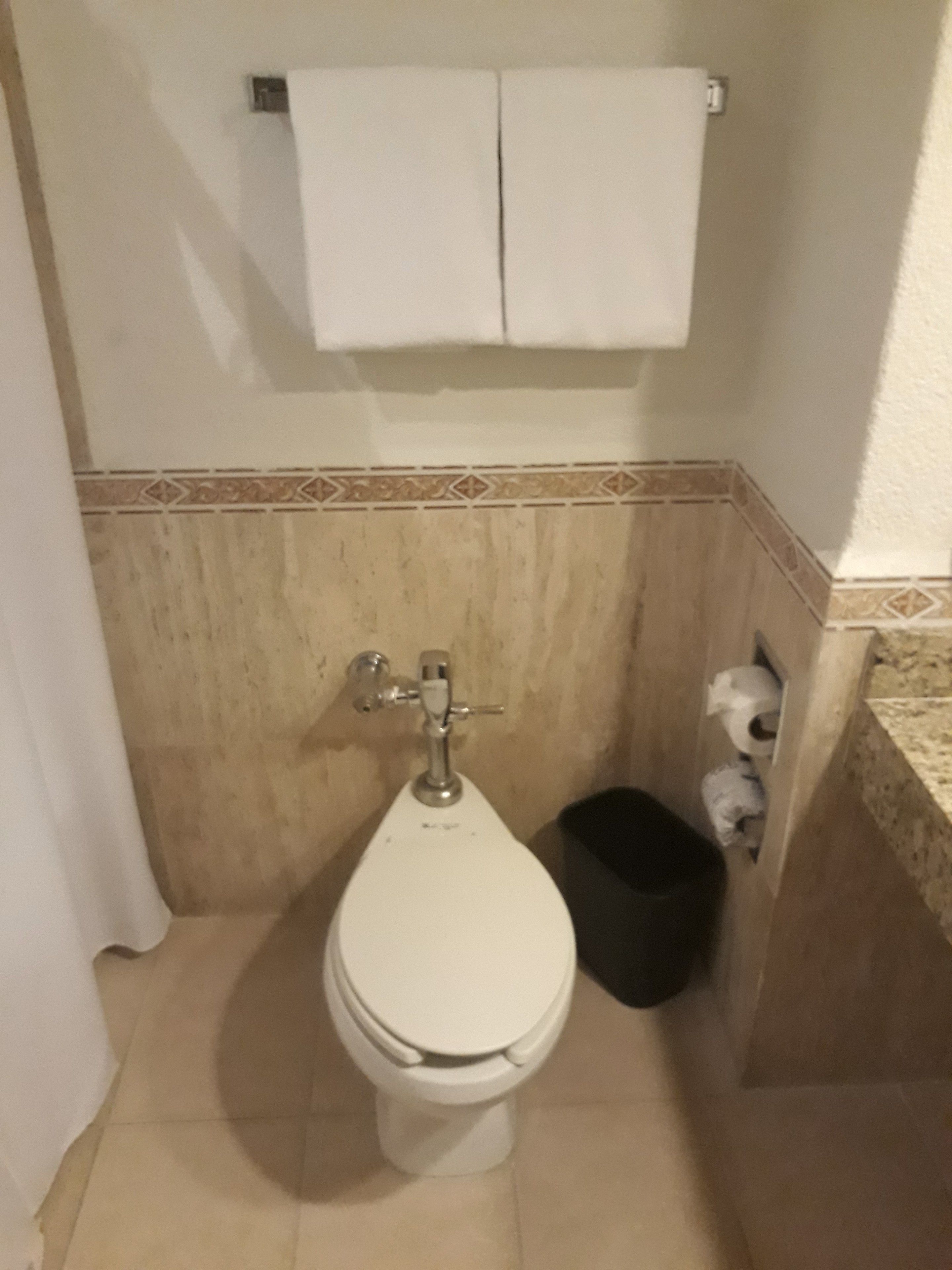 A small bathroom with a toilet, a towel rack above it, and a trash bin to the side.