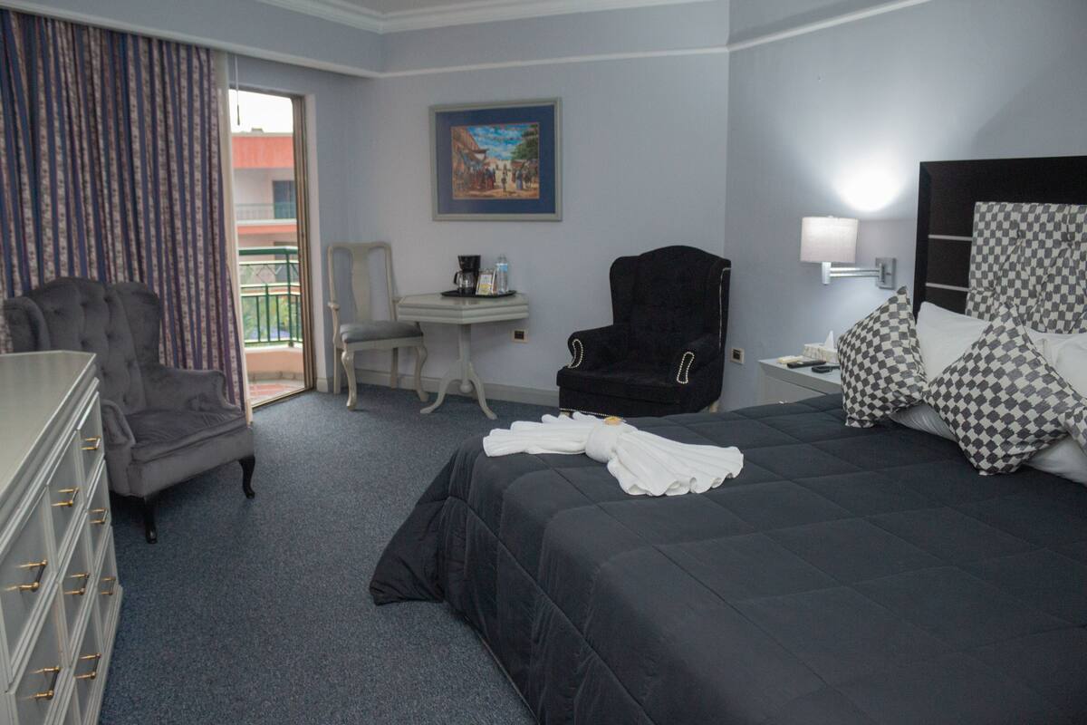 A hotel room with a large bed, a desk with a chair, a dresser, and a view of the outside balcony.