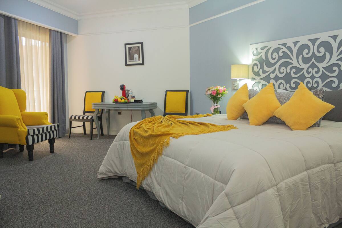 A hotel room with a bed, two chairs, a table with fruits, a vase with flowers, and a lamp.