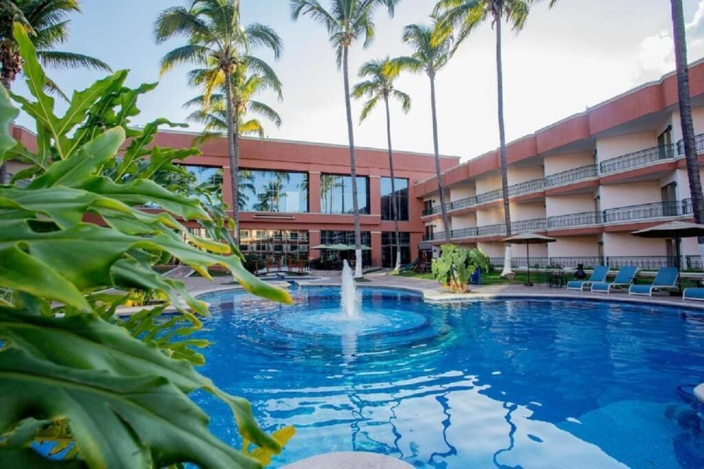 Une piscine avec une fontaine, entourée de palmiers et un immeuble à plusieurs étages doté de balcons.