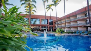 A swimming pool with a fountain, surrounded by palm trees and a multi-story building with balconies.