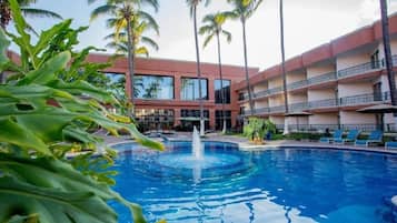 Une piscine avec une fontaine, entourée de palmiers et un immeuble à plusieurs étages doté de balcons.