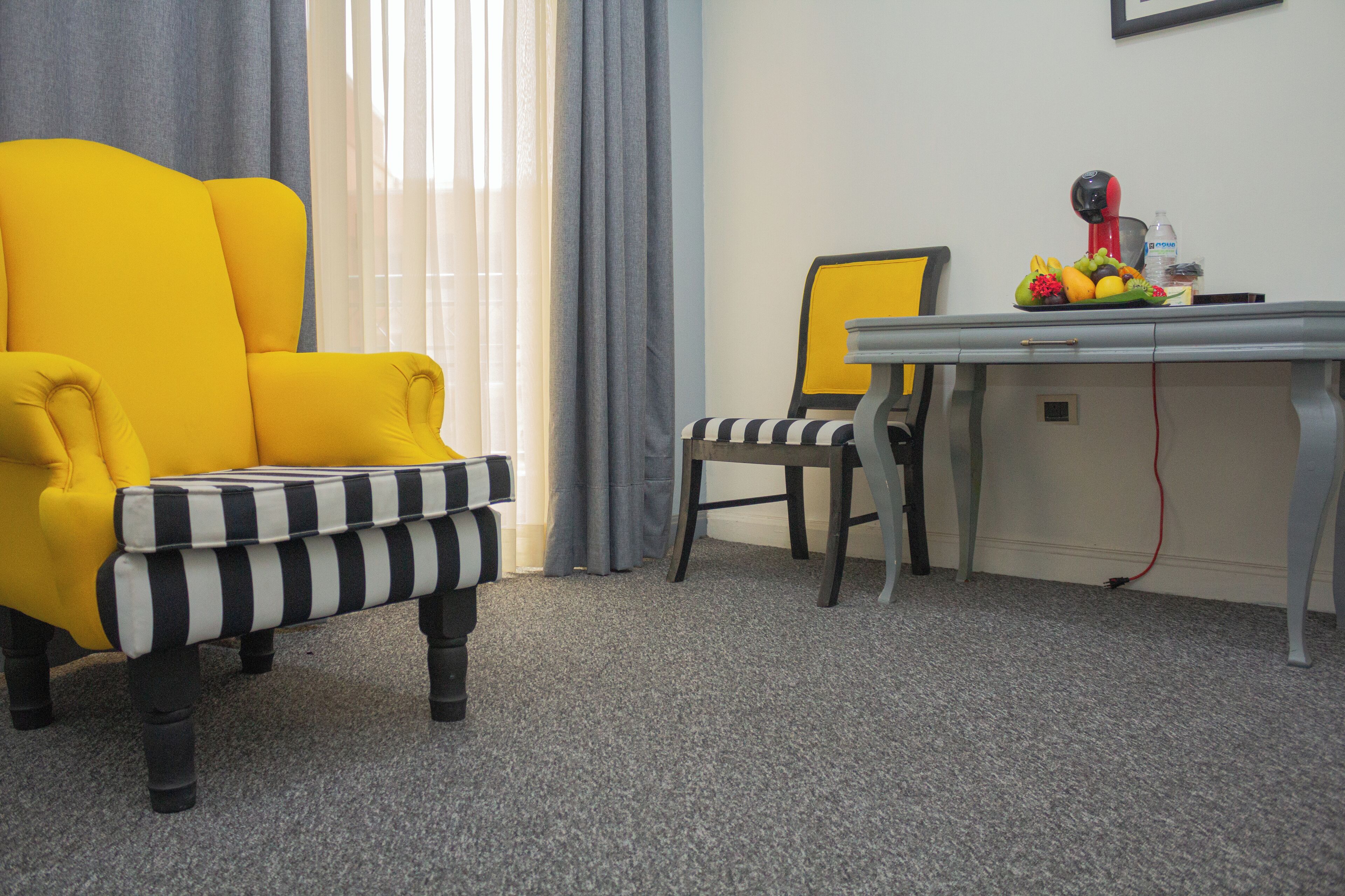 Een kamer met een gele fauteuil, een gestreept voetenbankje, een bureau met een stoel en een fruitschaal op het bureau.
