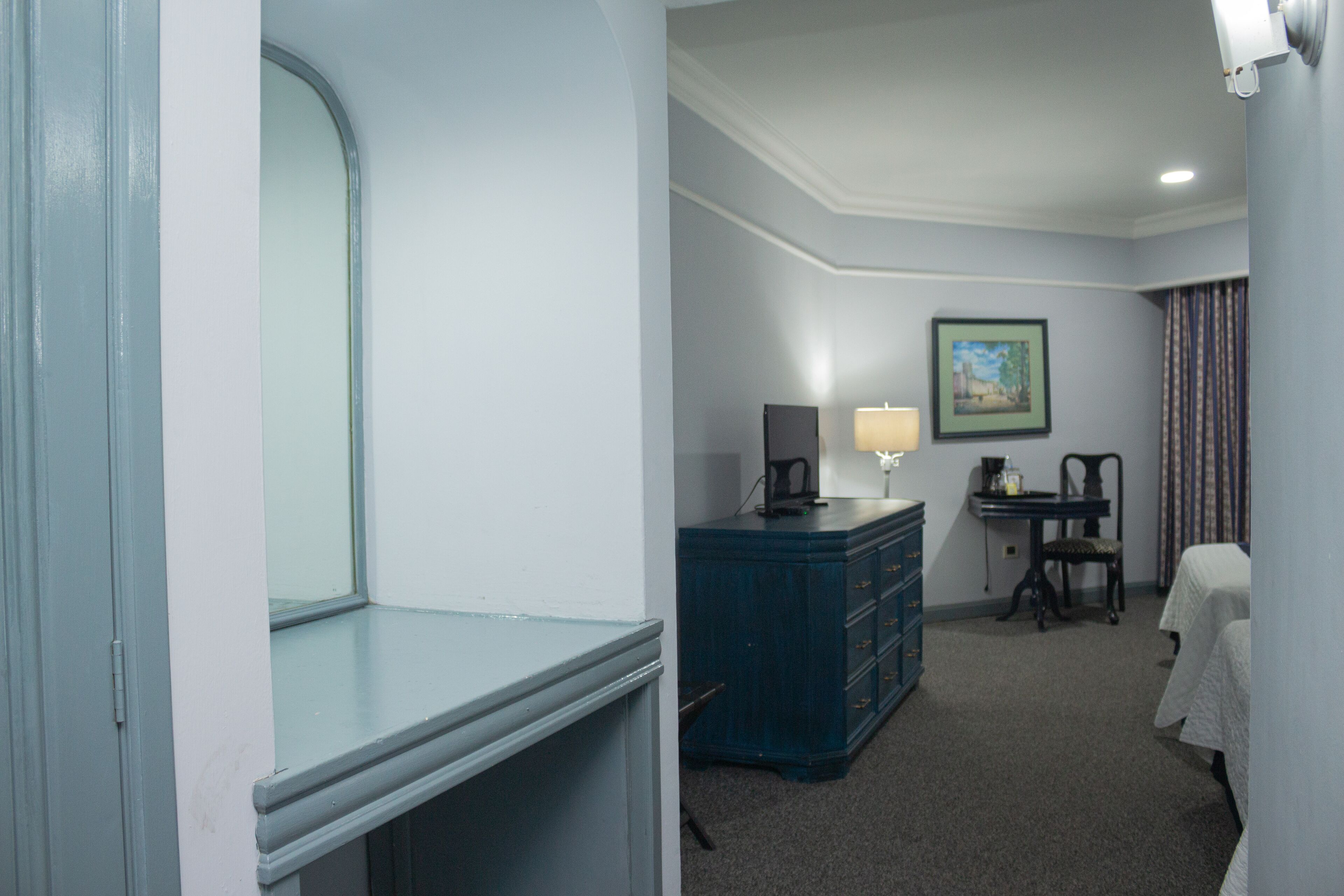 A hotel room with a large window, a desk with a lamp, a dresser, a chair, and a bed.