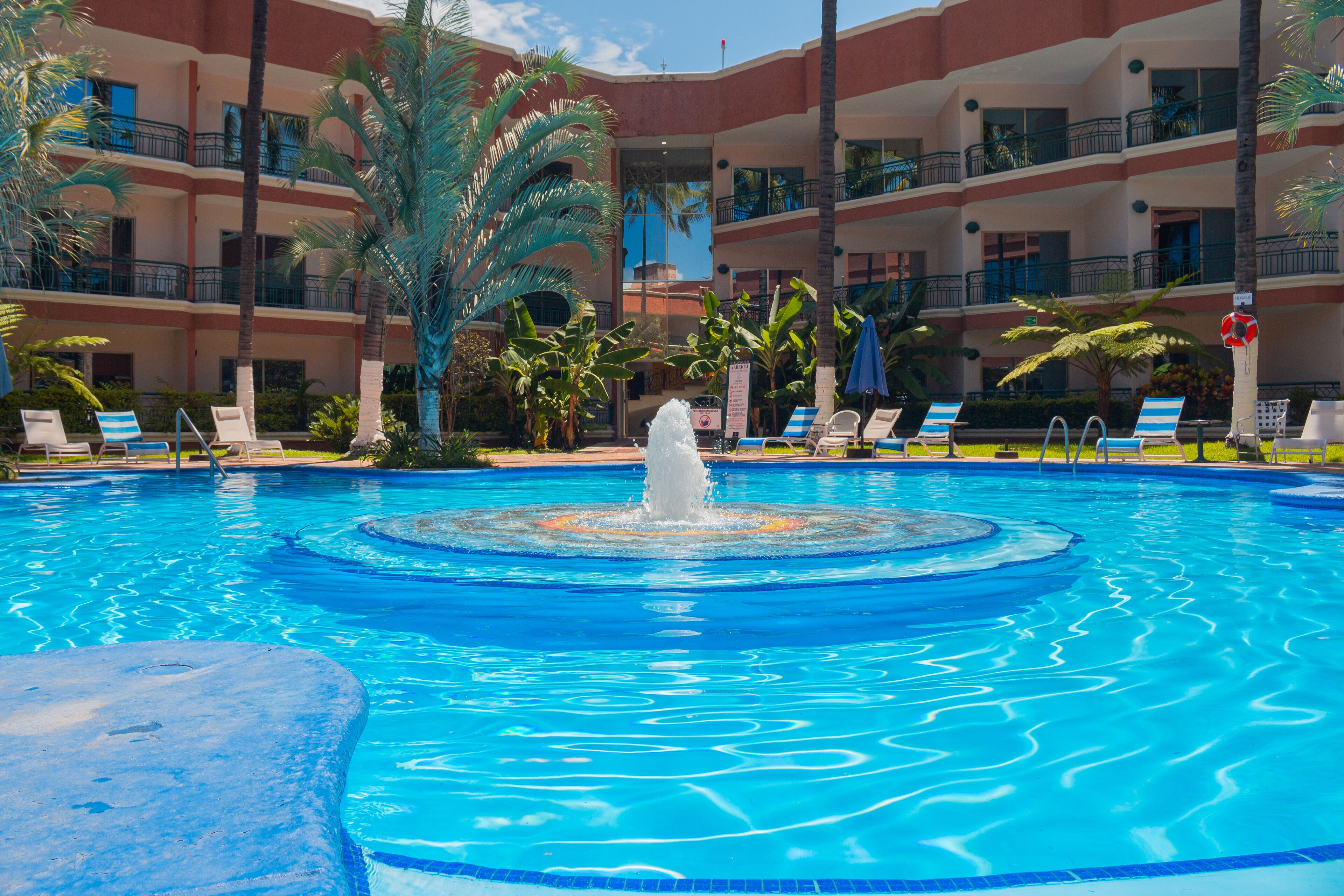 Una piscina con una fuente central, rodeada de tumbonas y palmeras.
