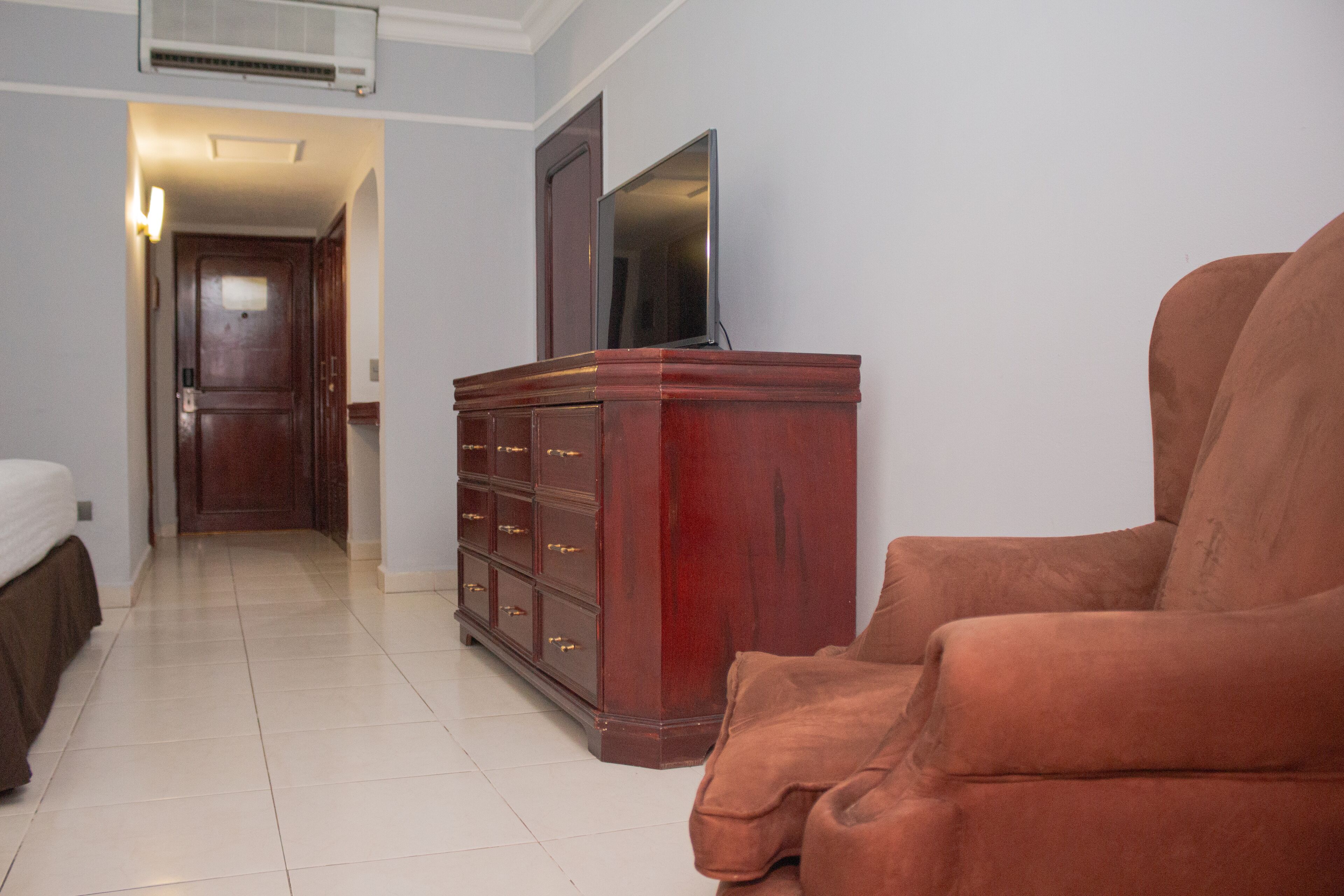 A hotel room with a bed, a wooden dresser, a television, and a brown armchair.