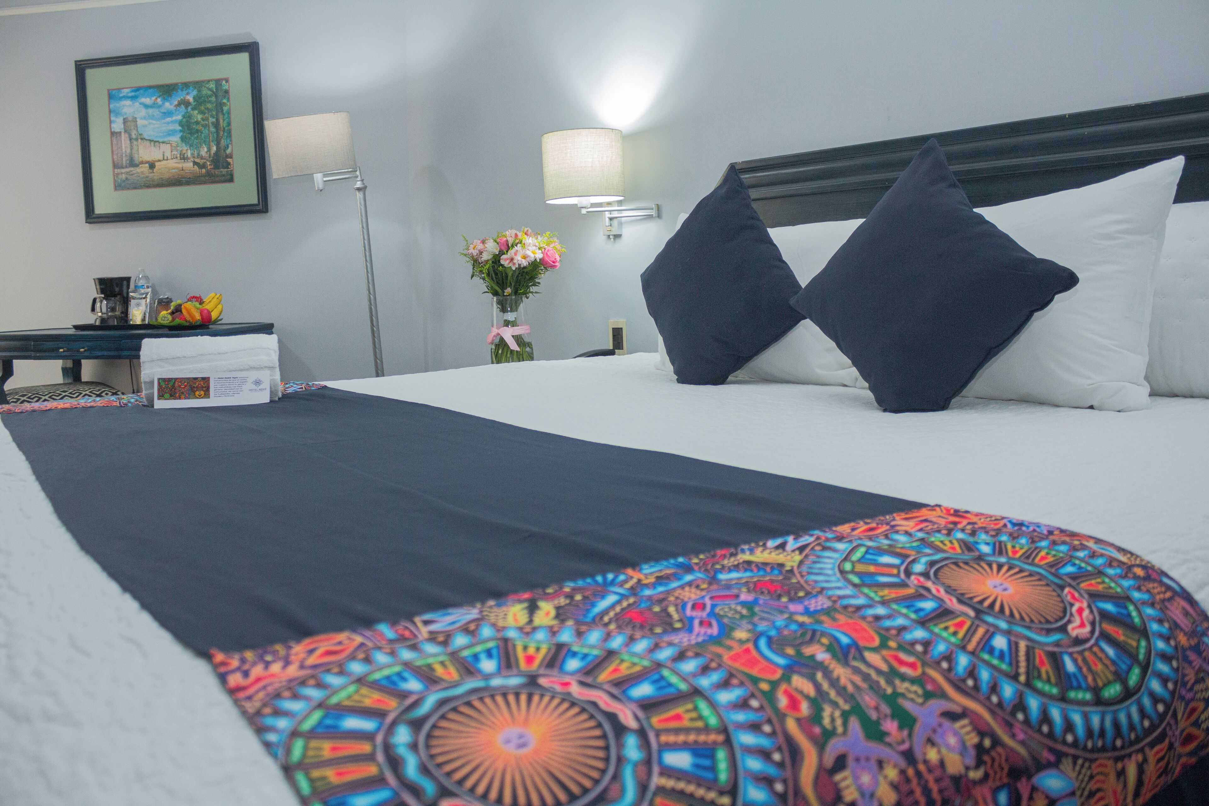 Una camera d'albergo con un letto rifatto, un comodino con una lampada, un quadro appeso alla parete e un vaso con fiori.