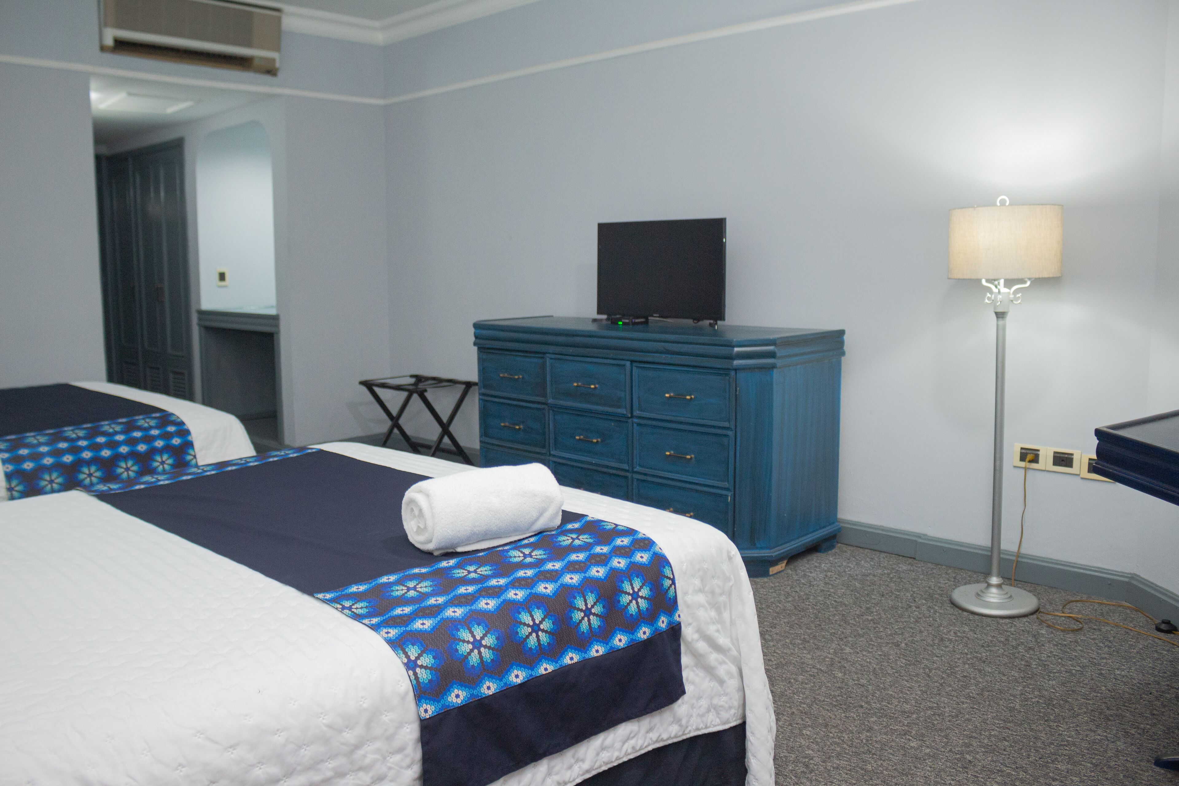 Una habitación de hotel con dos camas, un aparador azul, un televisor y una lámpara de pie.