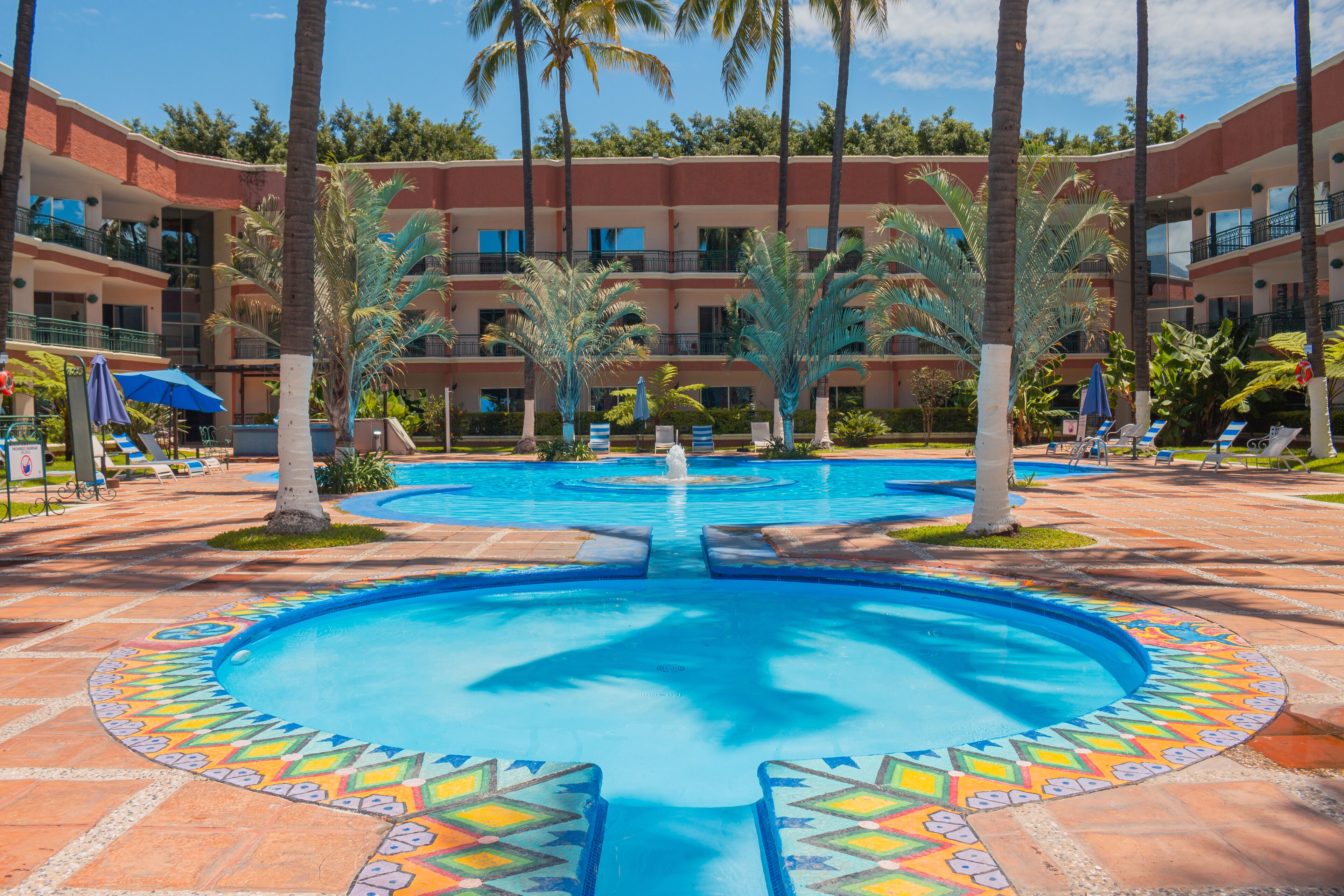 Un espace piscine d’hôtel comprenant plusieurs piscines, des palmiers et des chaises longues.