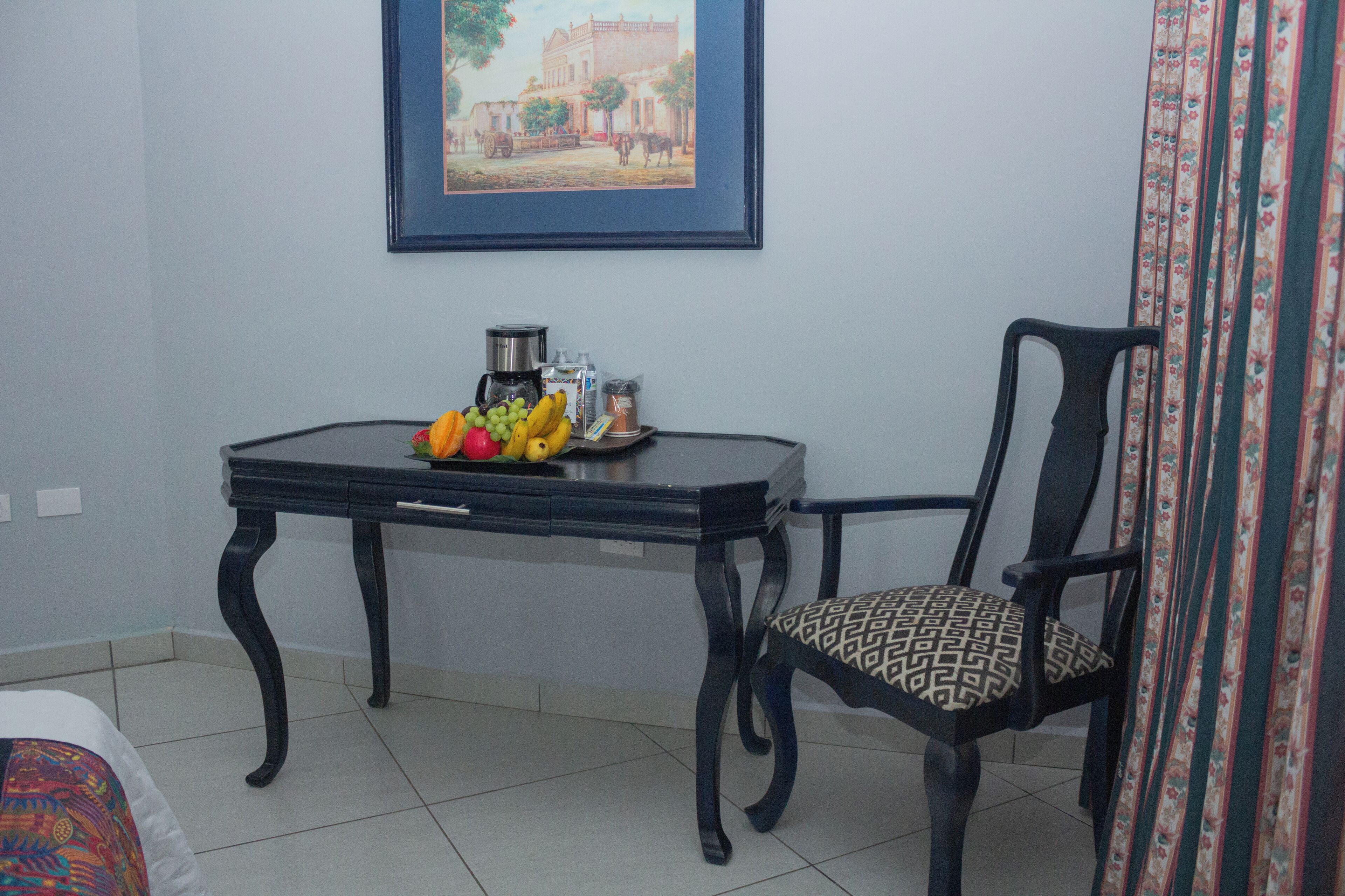 A room with a dining table set with fruits and a coffee maker, a chair with a patterned cushion, and a framed picture on the wall.