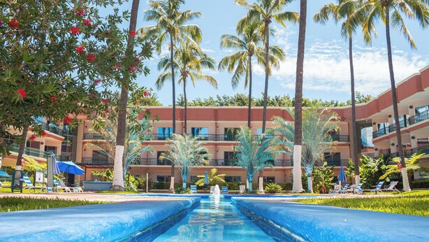 A swimming pool with a fountain, surrounded by palm trees and lounge chairs, with a hotel building in the background.