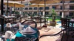 Un área de comedor al aire libre con mesas y sillas, sombrillas y una piscina al fondo.