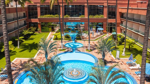 Un hotel con piscina central, palmeras y fachada de edificio roja.