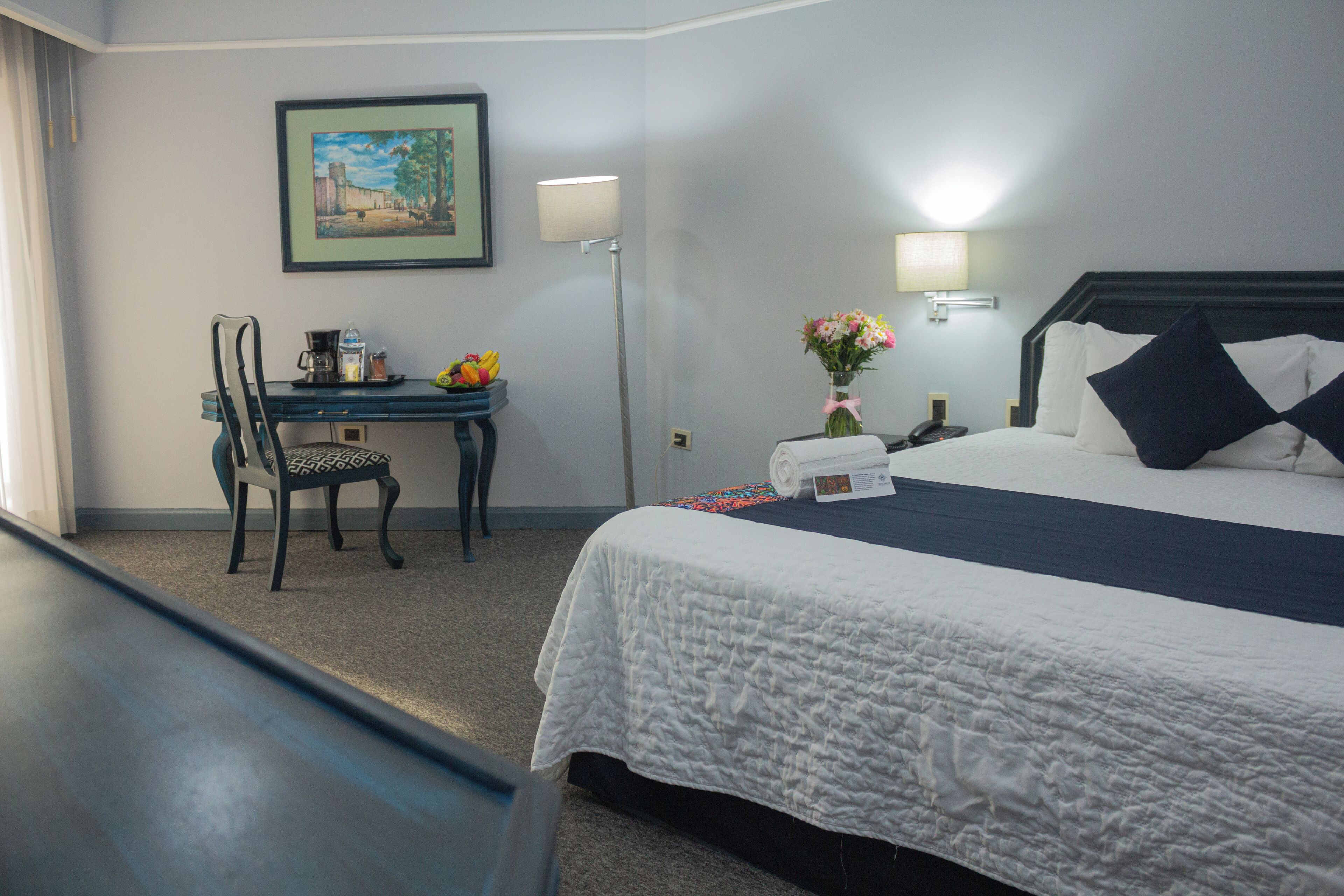 A hotel room with a bed, a desk with a chair, a lamp, a vase with flowers, and a framed picture on the wall.