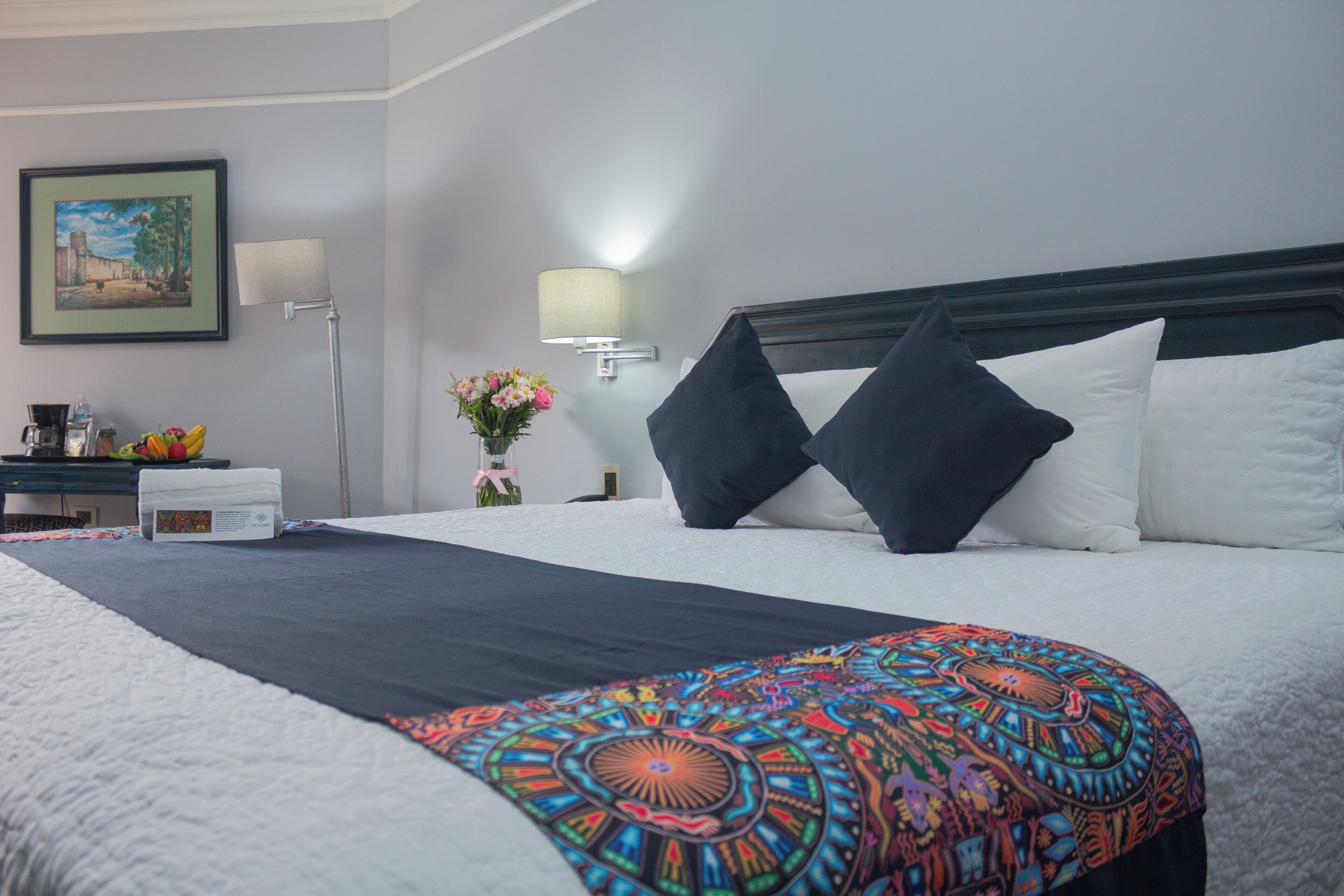 Una camera d'albergo con un letto grande, un comodino con una lampada, un quadro appeso al muro e un vaso con fiori.
