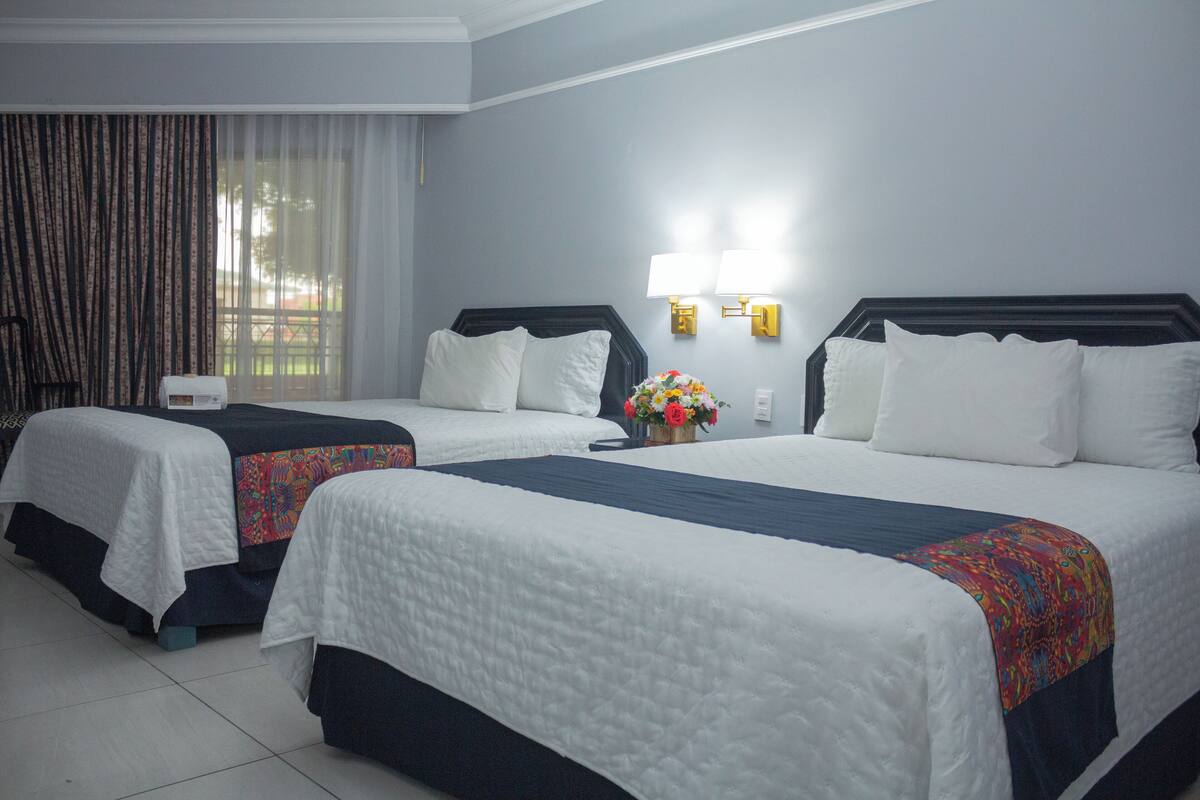 A hotel room with two beds, a large window with curtains, and a wall-mounted lamp.