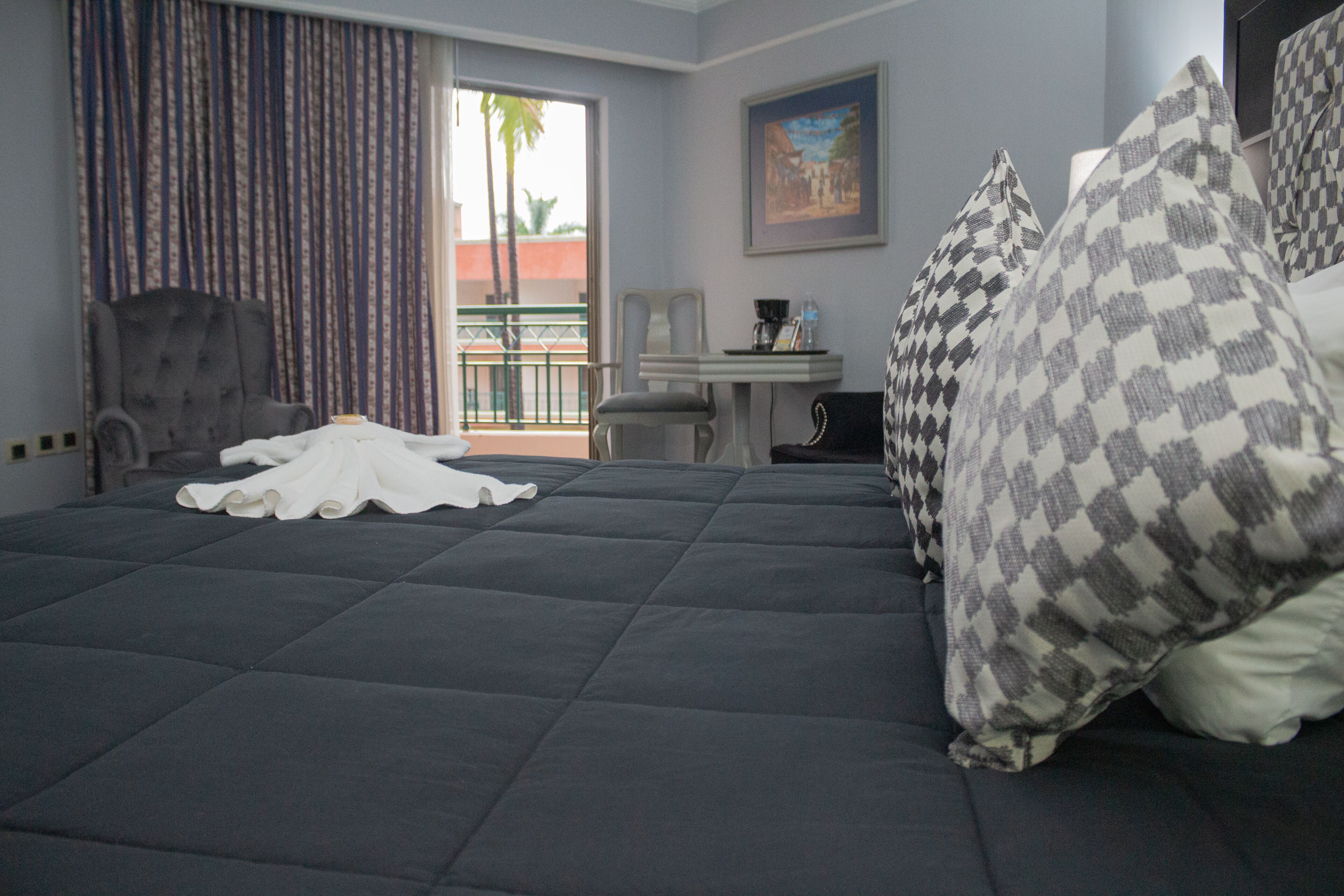 Un cuarto de hotel con una cama grande, un escritorio, una silla, un balcón con vista a palmeras y un cuadro en la pared.