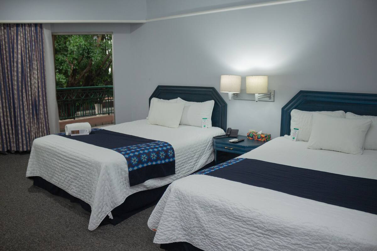 A hotel room with two beds, a nightstand, a lamp, and a window with a view of trees.