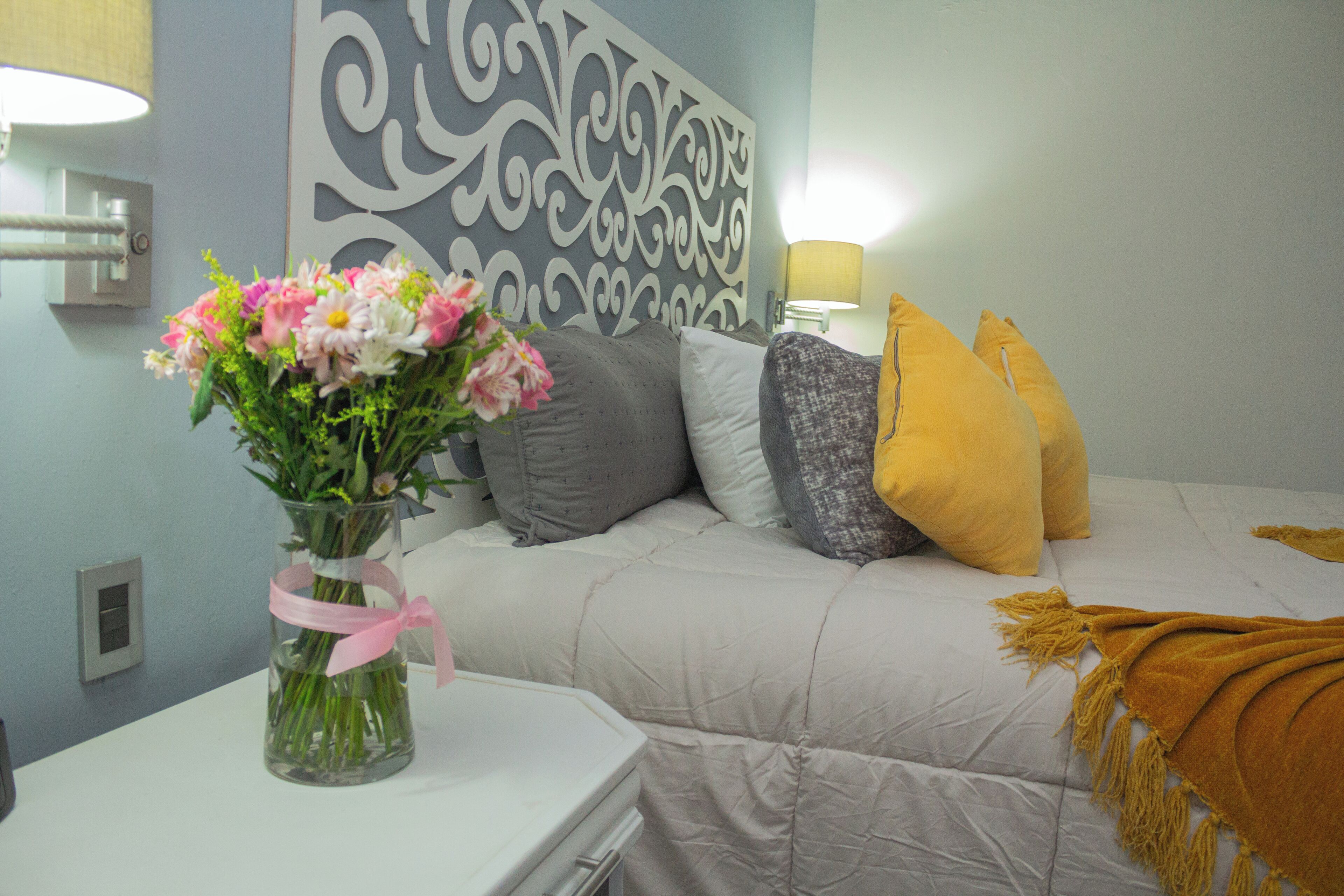 Un dormitorio con cama, almohadas, un jarrón con flores y una mesita de noche.