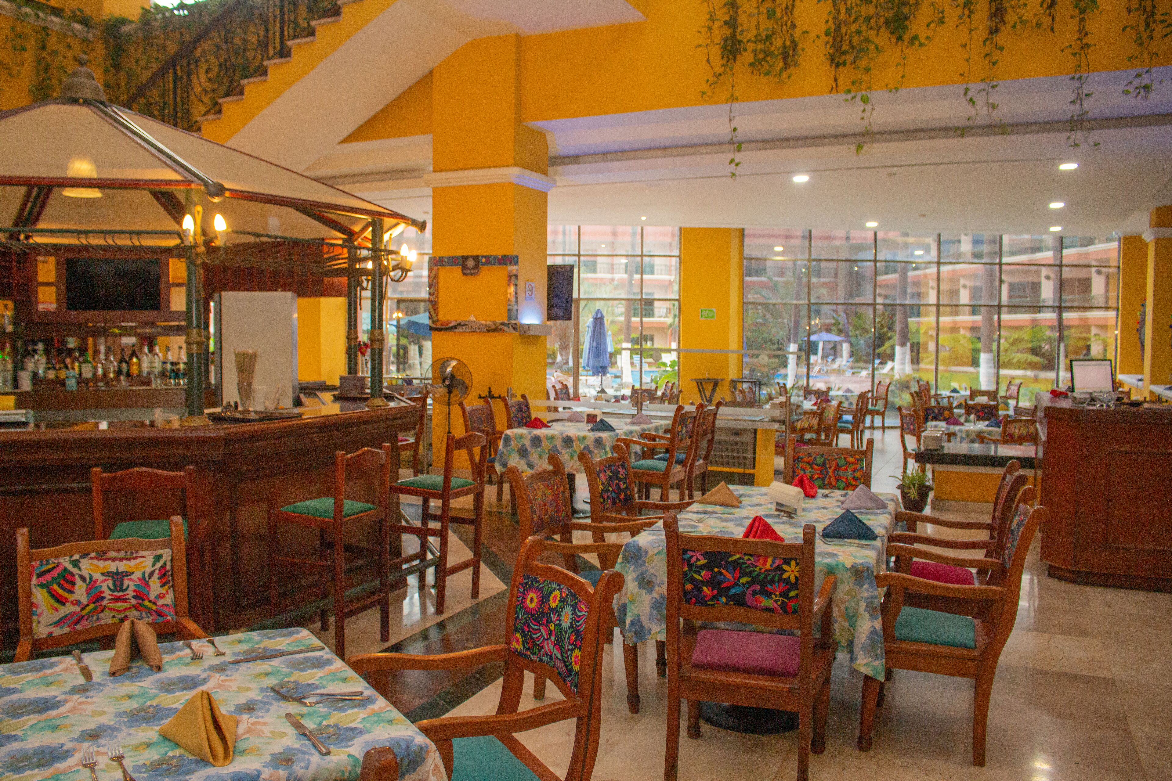 Un restaurante con varias mesas y sillas, una zona de bar y grandes ventanales que ofrecen vistas al exterior.