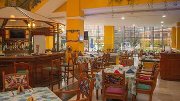 Un restaurante con varias mesas y sillas, una zona de bar y grandes ventanales que ofrecen vistas al exterior.