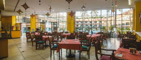 A spacious indoor dining area with multiple tables and chairs, large windows offering a view of a pool and buildings.